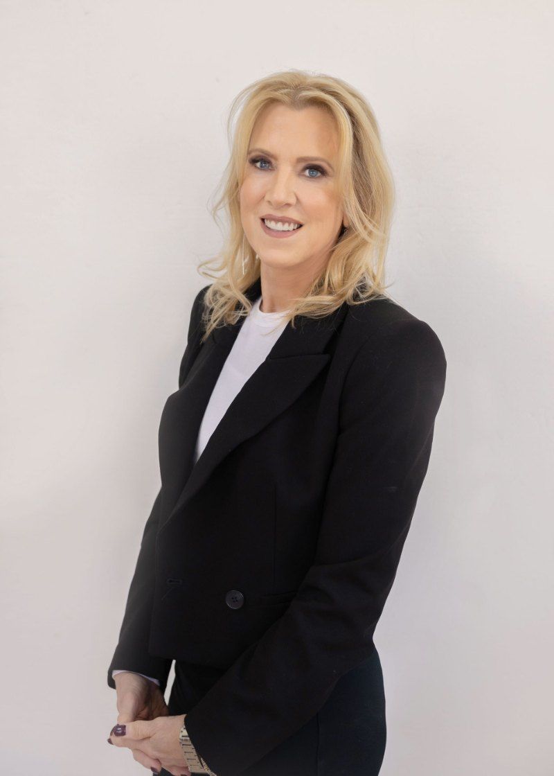 A woman in a black jacket and white shirt is standing in front of a white wall.