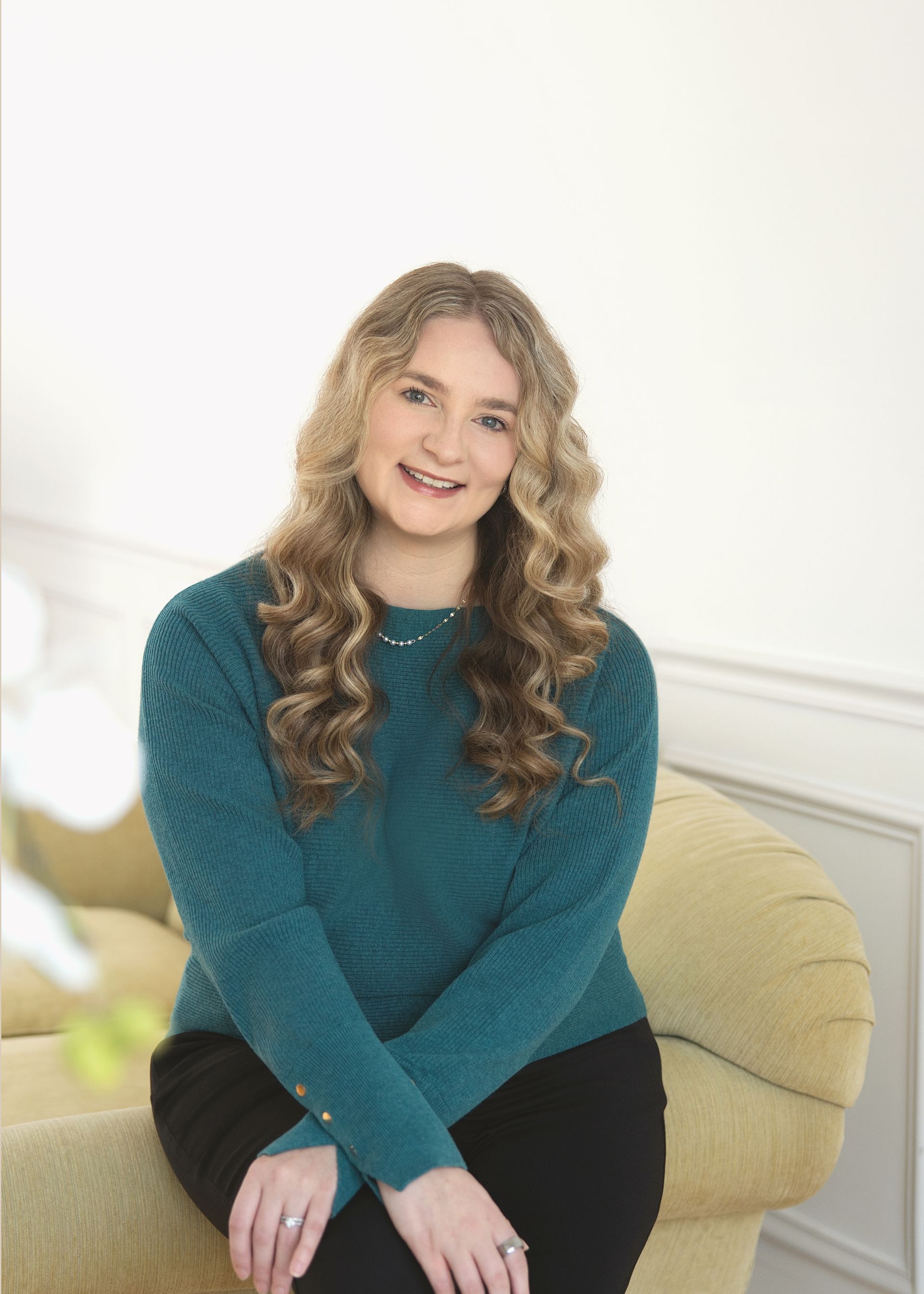 Woman with curly blonde hair sitting on a yellow chair, wearing a teal sweater and black pants.