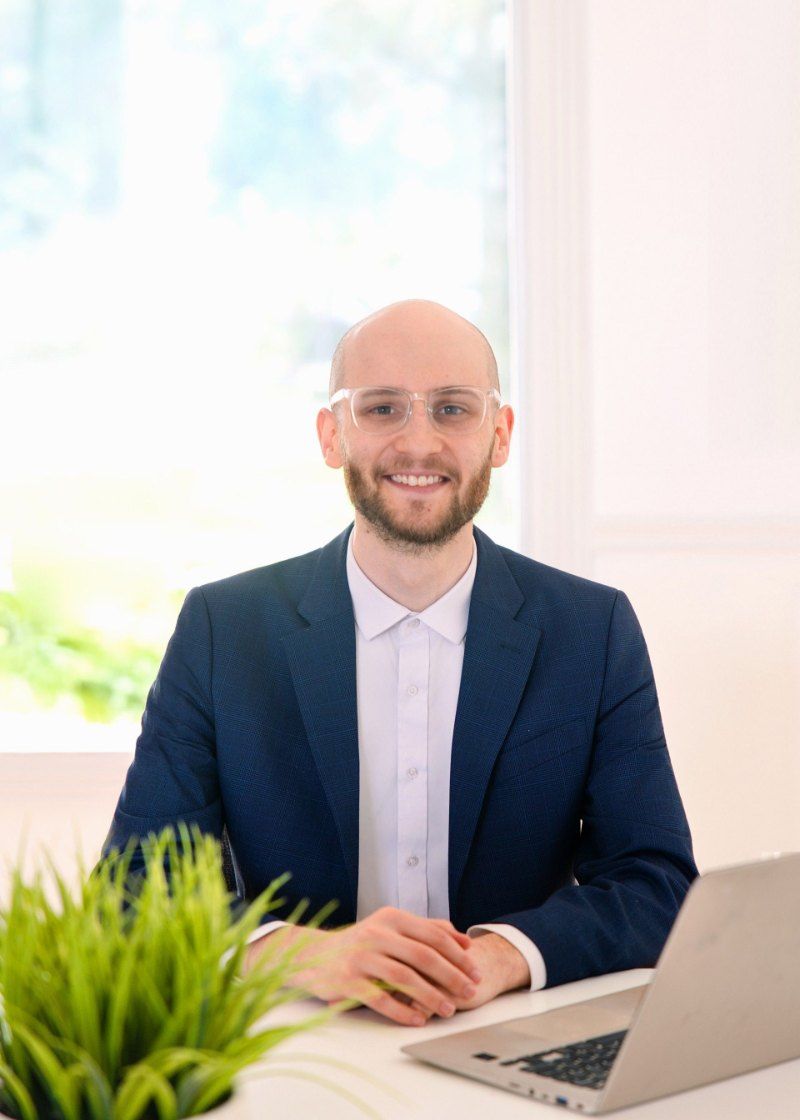 Man smiling at a desk with a laptop in a bright office, wearing a navy blazer and glasses