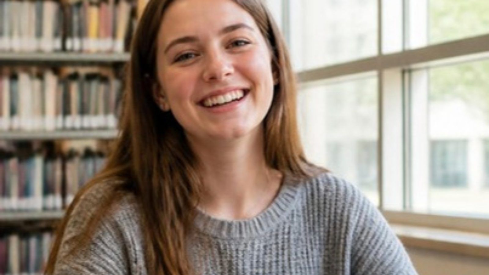 Smiling college student in a gray sweater by a sunny library window with bookshelves behind her