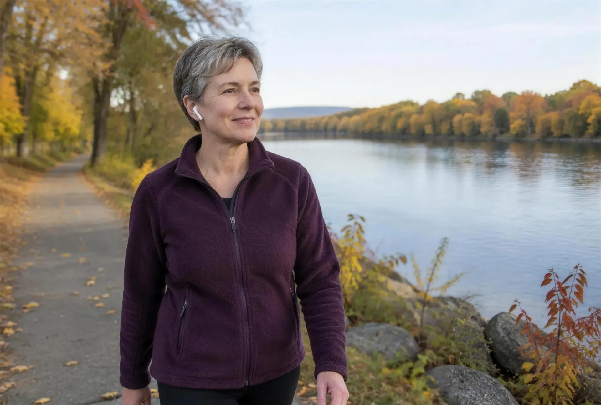 Woman with short gray hair smiling as she walks a riverside path on a fall morning