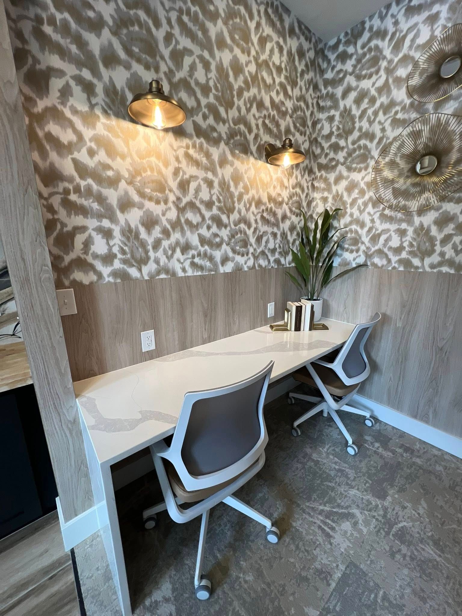 Office nook with white desk and two chairs, beige patterned wallpaper, and sconce lighting.