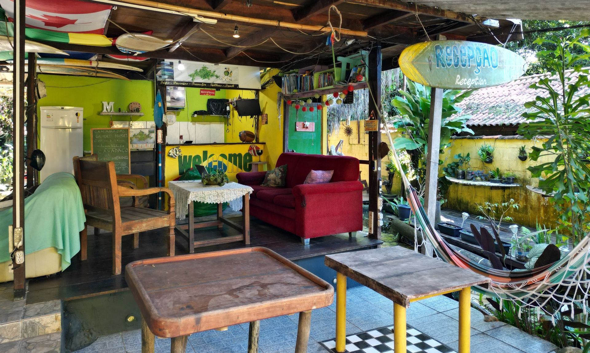 Rustic and cozy reception area in Ilha Grande, featuring a relaxed atmosphere with a red sofa, wooden tables, hammock, tropical plants, and colorful decorations, perfect for travelers seeking local hospitality and charm.