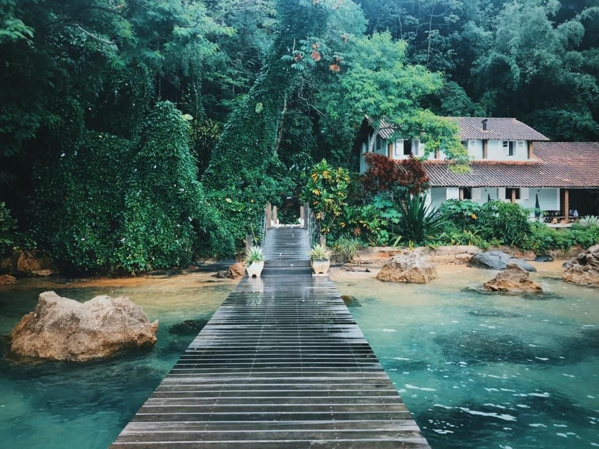 Picturesque view of a charming guesthouse surrounded by lush greenery and tropical plants, located in Ilha Grande. The scene features a wooden boardwalk leading over crystal-clear waters, creating a serene and inviting setting ideal for relaxation
