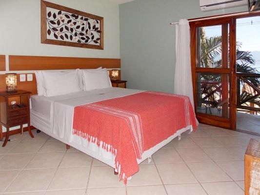 Comfortable suite at Pousada Caiçara in Vila do Abraão, Ilha Grande, featuring a neatly made bed with white linens and a red throw blanket, bedside tables with lamps, wall art, and a balcony with a view of tropical greenery, offering a cozy and inviting atmosphere