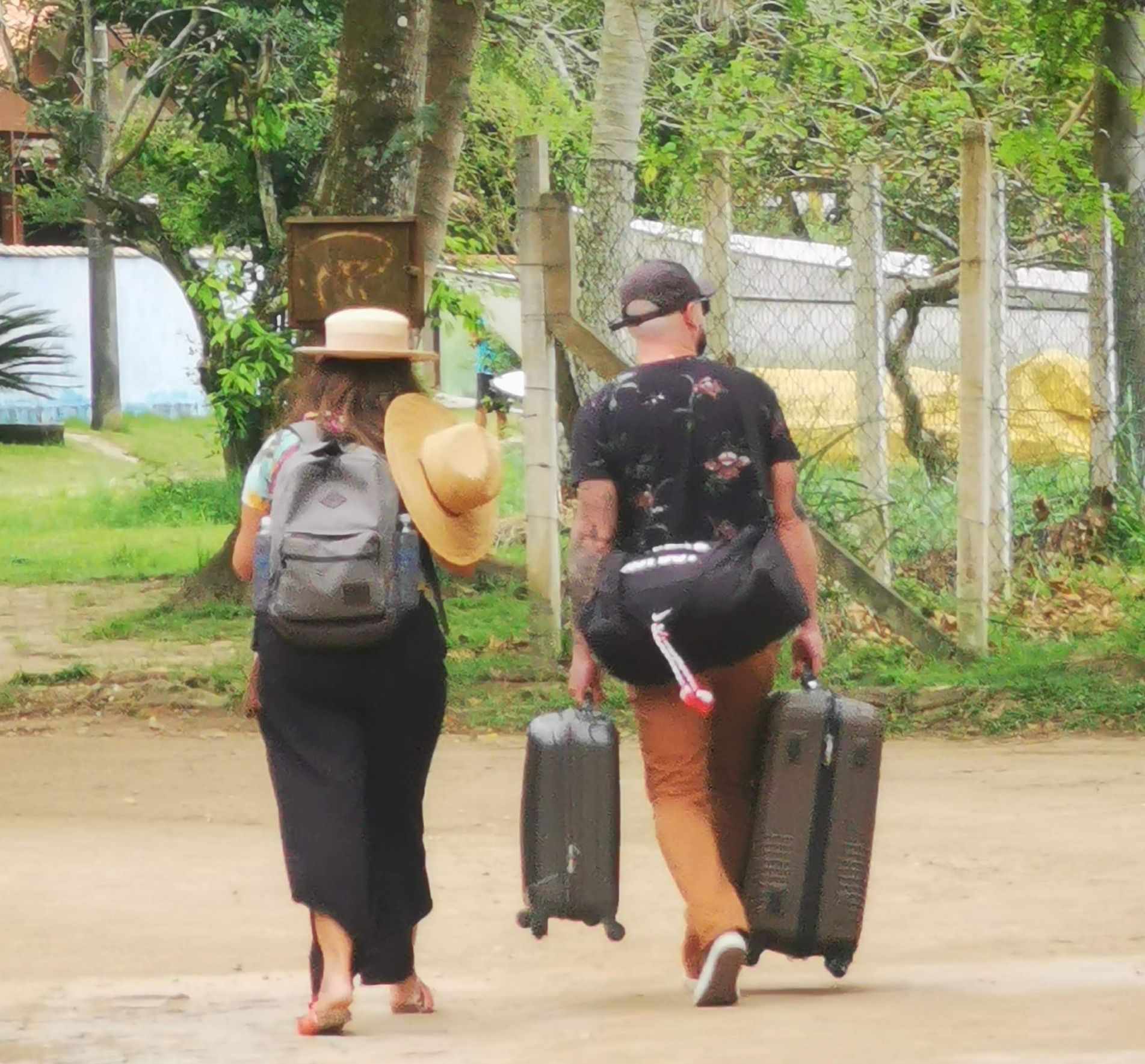 Um casal caminha por uma estrada de terra na Vila do Abraão, cada um carregando malas de rodinhas e bolsas grandes. A mulher usa um chapéu de palha e mochila, e o homem veste uma camisa estampada com uma mala de mão. Embora a imagem mostre turistas chegando para se hospedar, ela é usada de forma divertida para ilustrar o que não levar em uma caminhada na Ilha Grande, destacando a diferença entre o que se usa para viagens e o que é mais adequado para trilhas e aventuras na natureza.