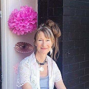 A woman is sitting in front of a black brick wall with pink pom poms hanging on the wall.