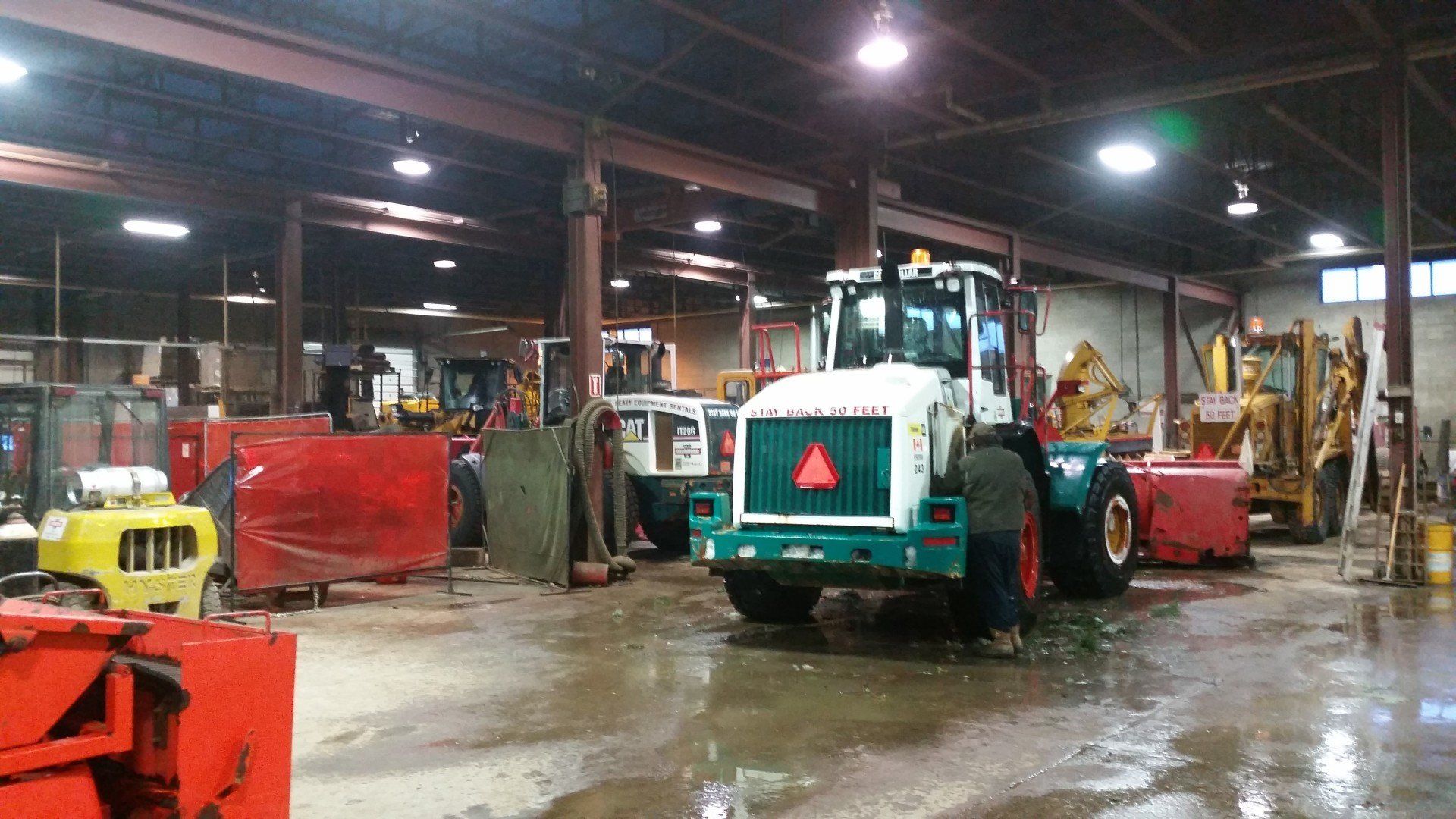 Large equipment indoors, mostly white and teal, in a workshop. Includes tractors, loaders, and snow plows. Wet floor.