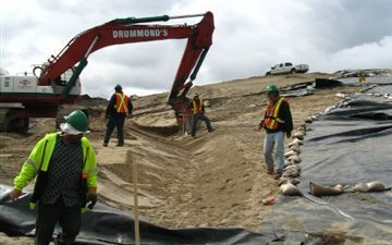Construction crew works on a hillside with an excavator. Black tarp and protective gear are visible.