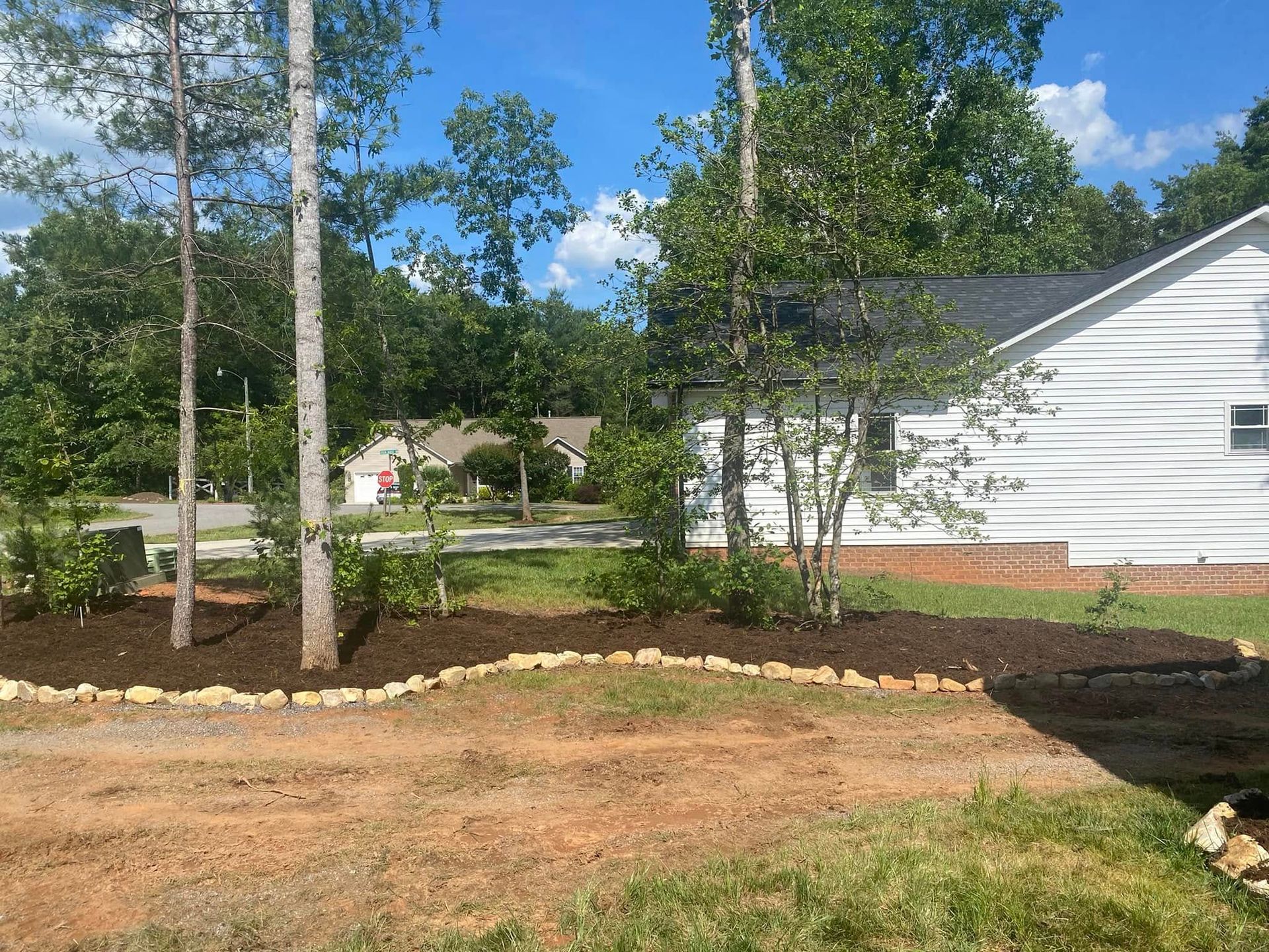 A freshly mulched garden bed with a stone border in front of a white house, surrounded by trees and a dirt yard.