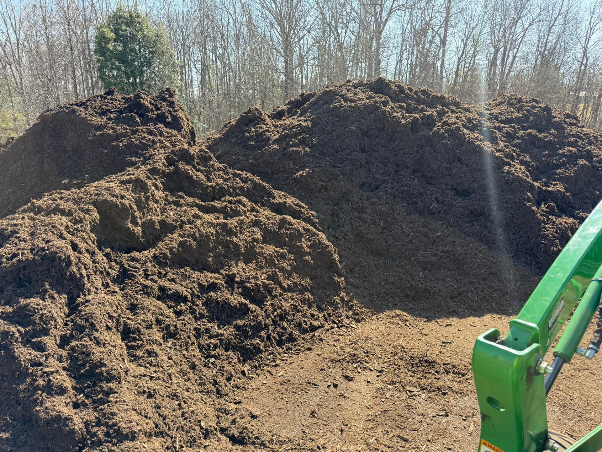 Two large piles of dark brown mulch sit in a field on a sunny day, with a piece of green farm equipment in the foreground.
