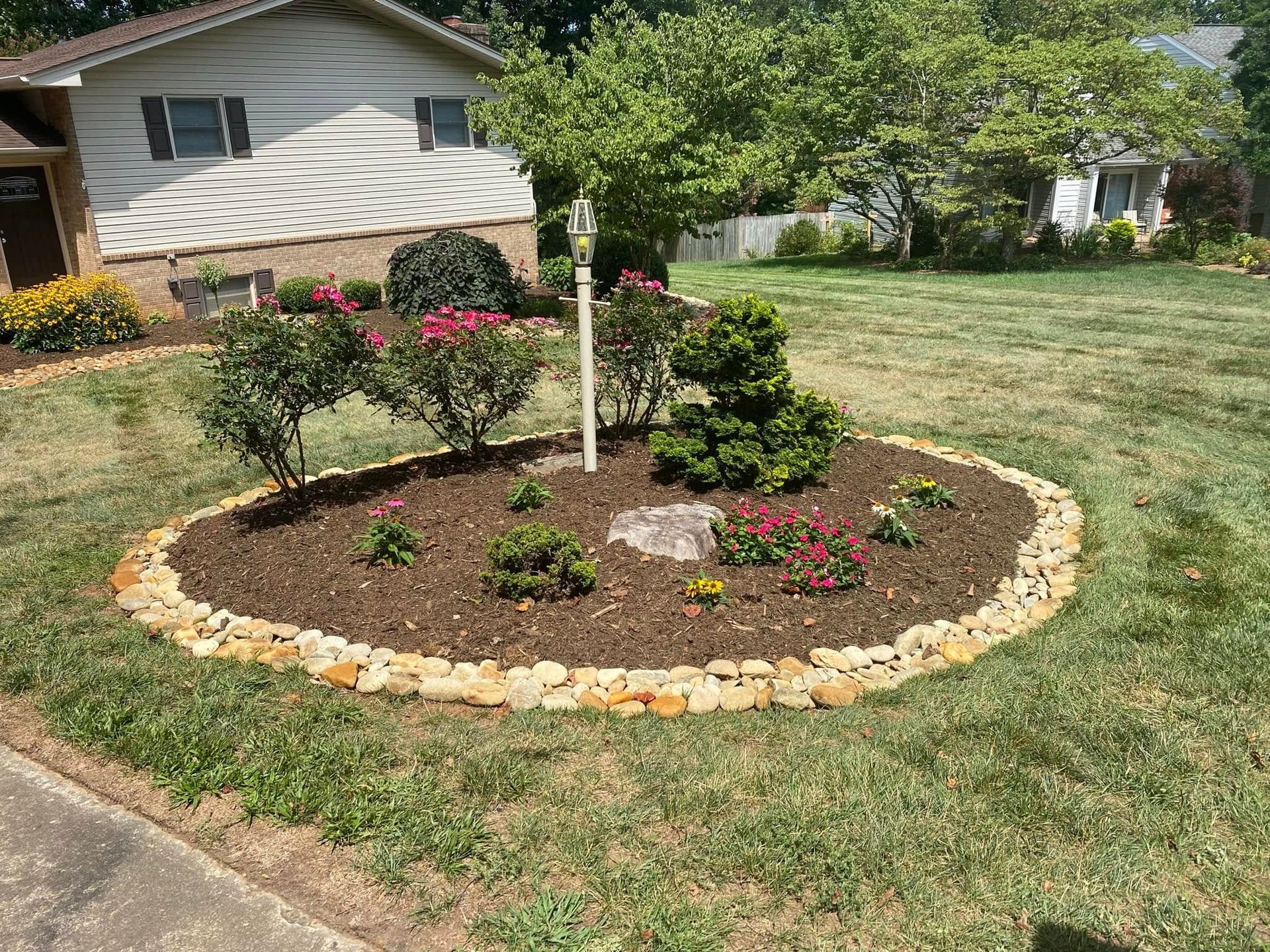 A circular garden bed with mulch, small bushes, and stone edging in a grassy front yard of a suburban home.