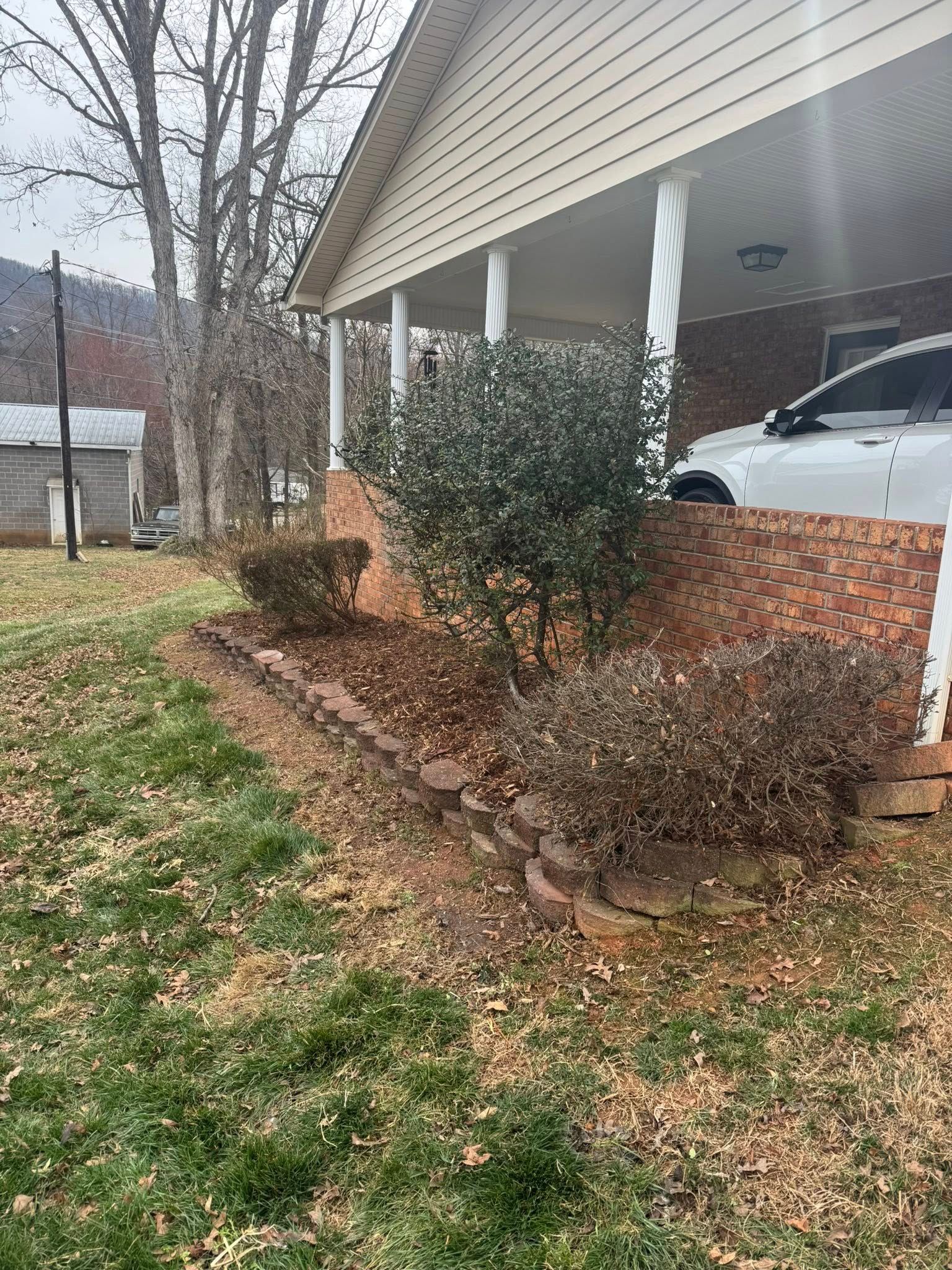 A garden bed with brown mulch and shrubs in front of a brick porch, with a parked white vehicle and trees in the background.