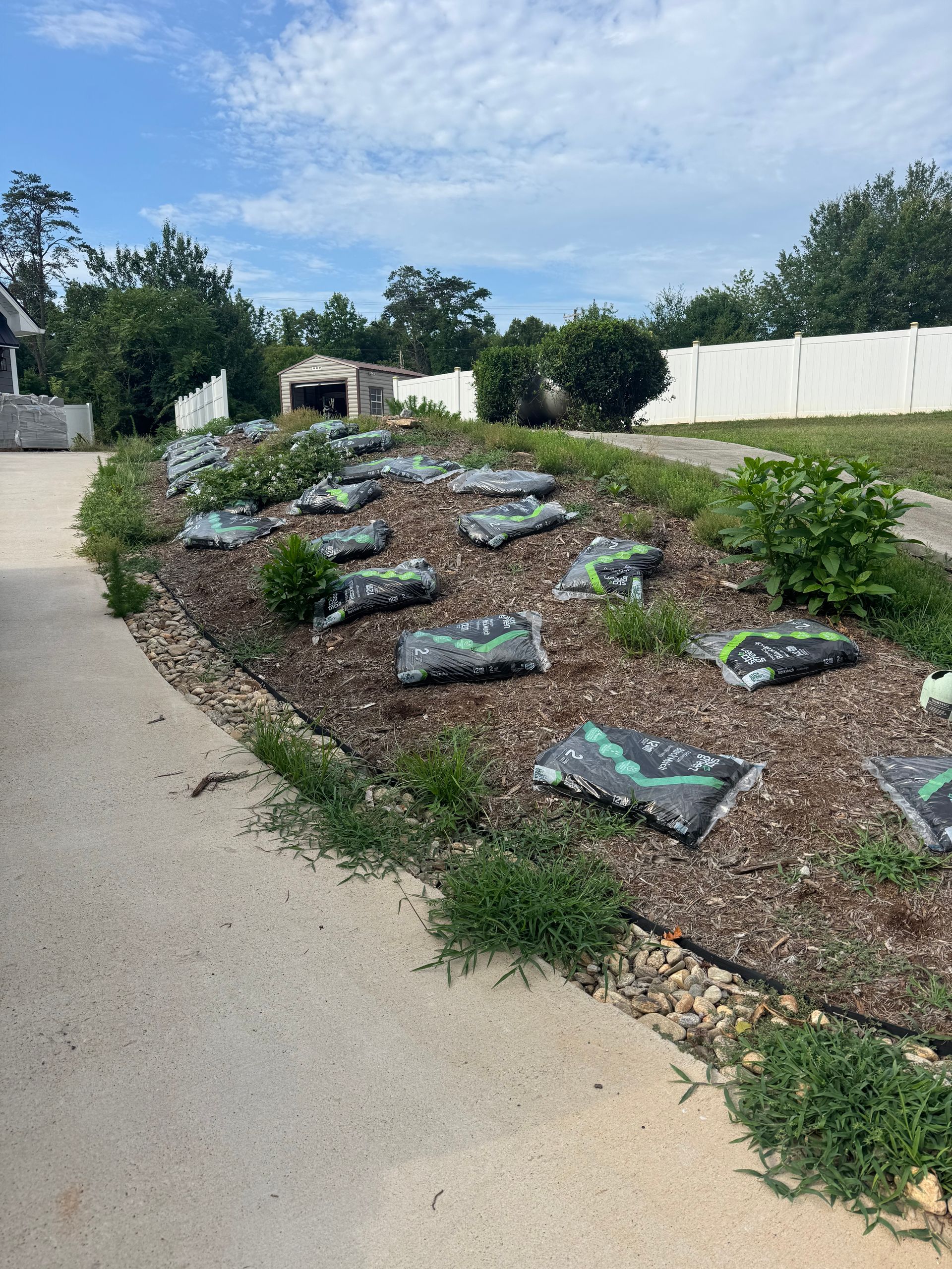 Mulched roadside planting bed with small green plants beside a concrete path and white fence