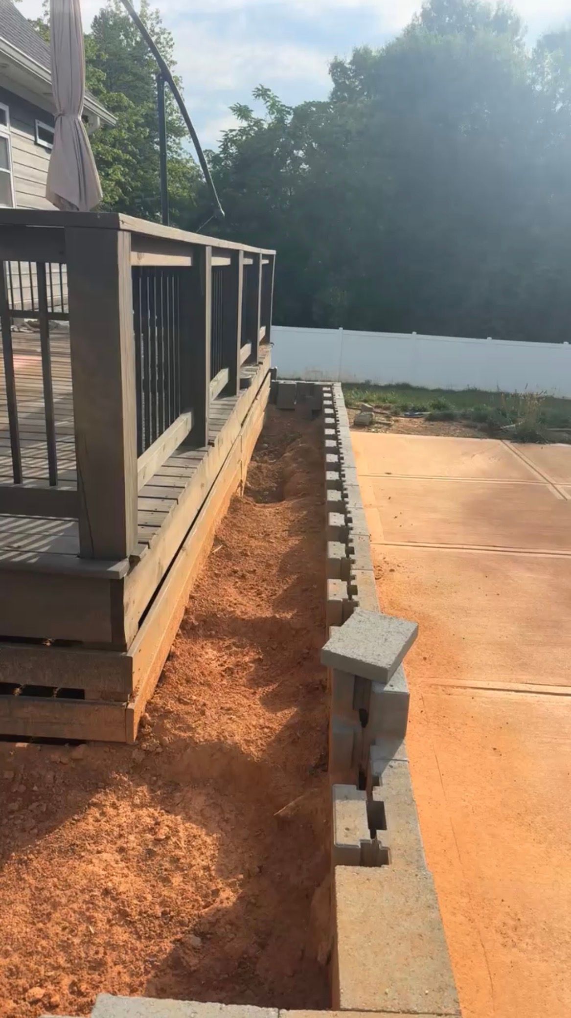 Wooden deck beside a narrow trench with cinder blocks, next to a concrete patio and white wall outdoors