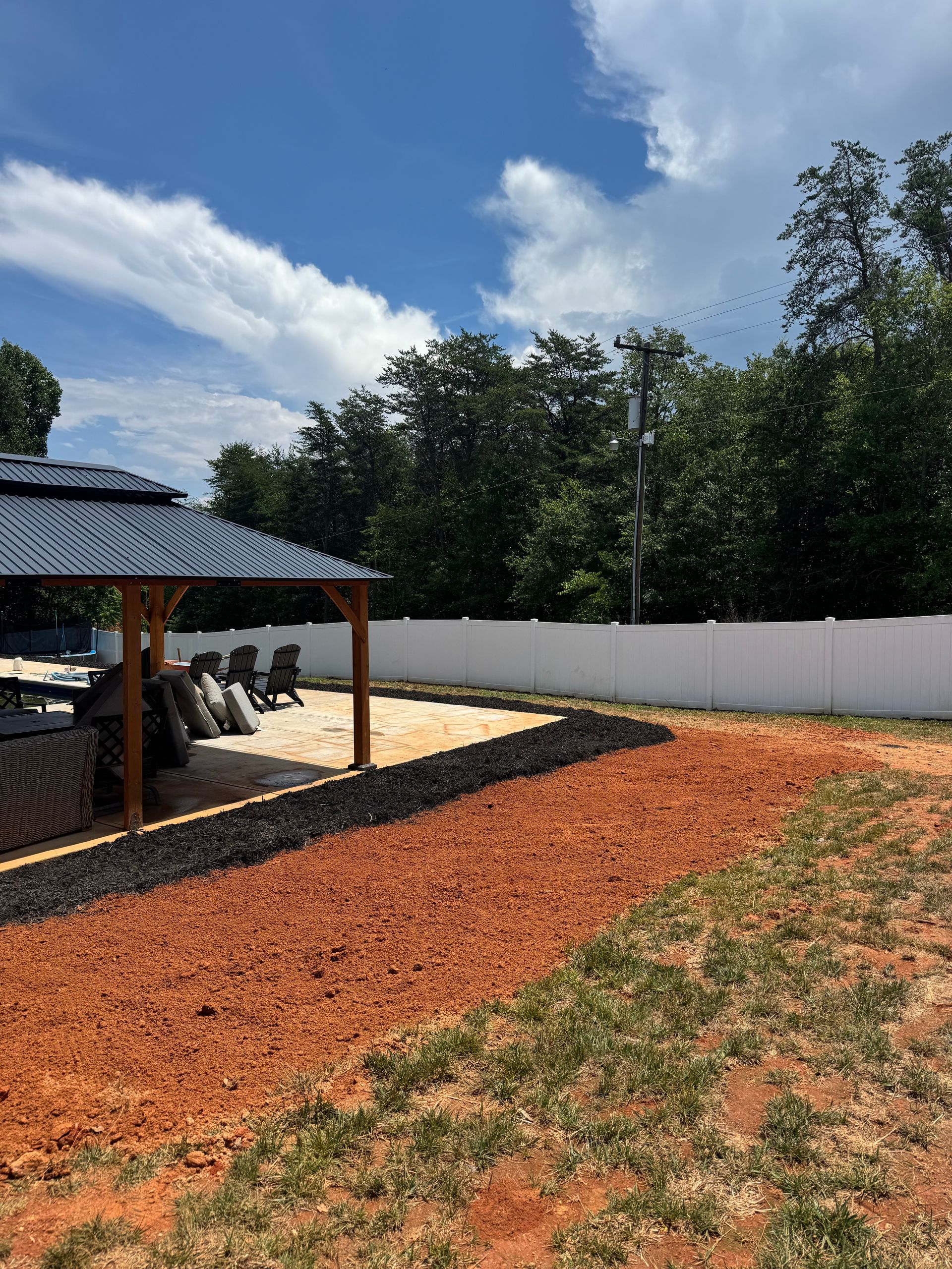 Backyard with red mulch, white fence, gravel path, and covered carport under a blue sky
