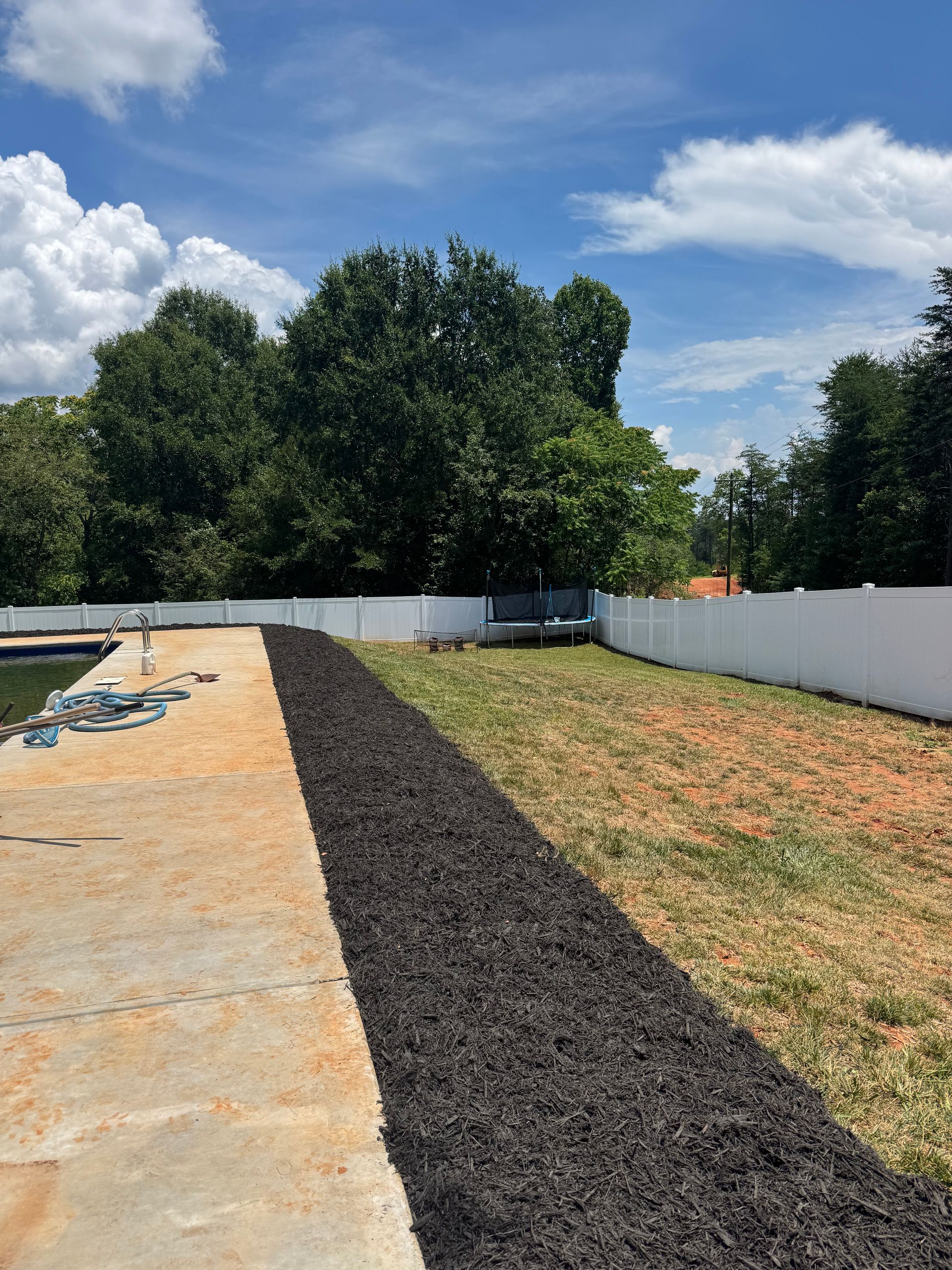 Gravel strip beside a concrete slab and white fence in a sunny backyard with trees and clouds.