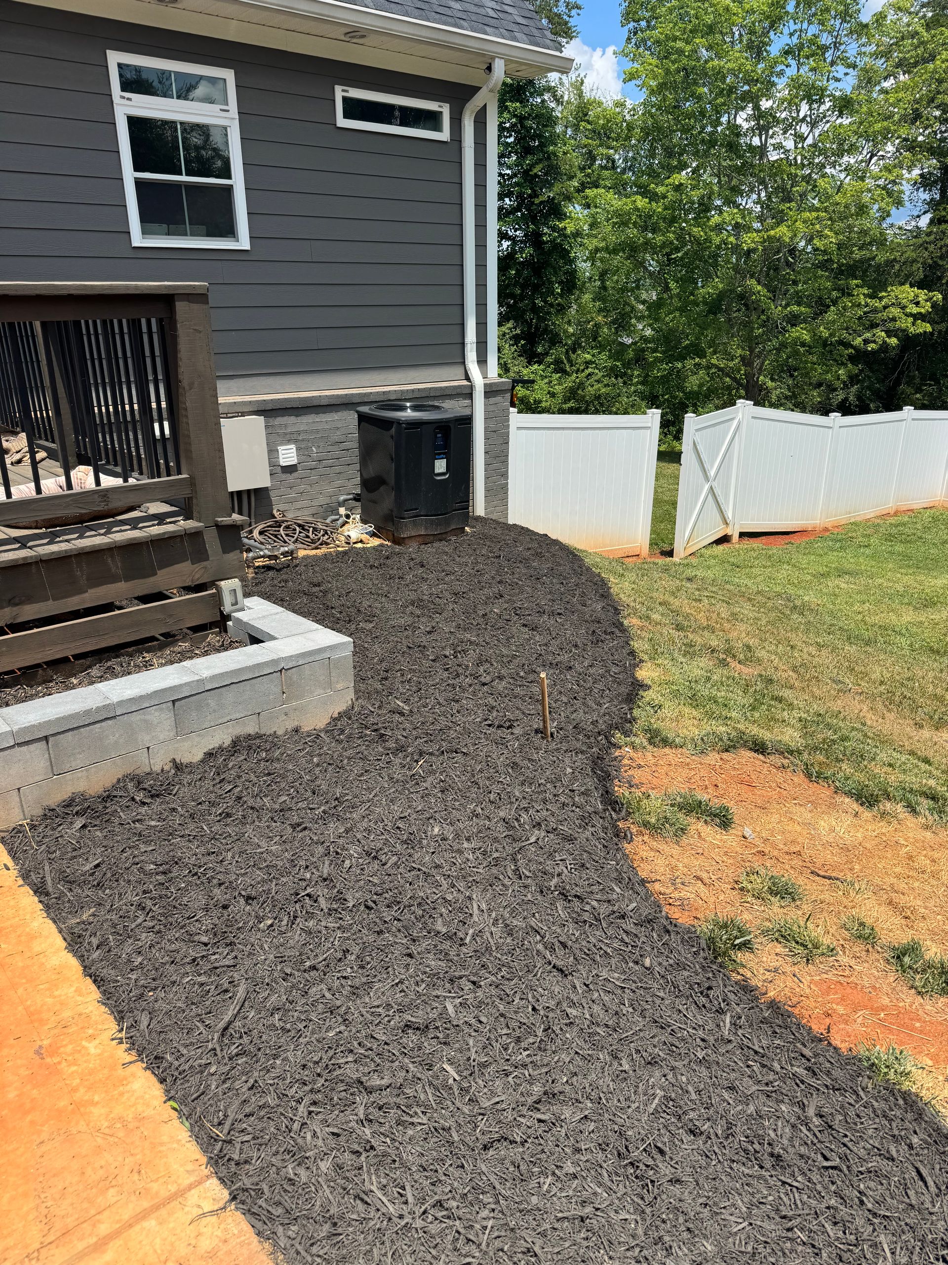 Dark gravel path beside a gray house and wooden porch, bordered by white fencing and grass.