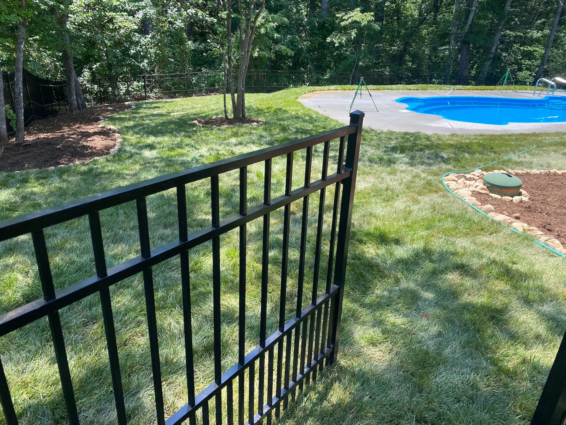 Black metal gate opening to a backyard with a swimming pool, lawn, and surrounding trees.