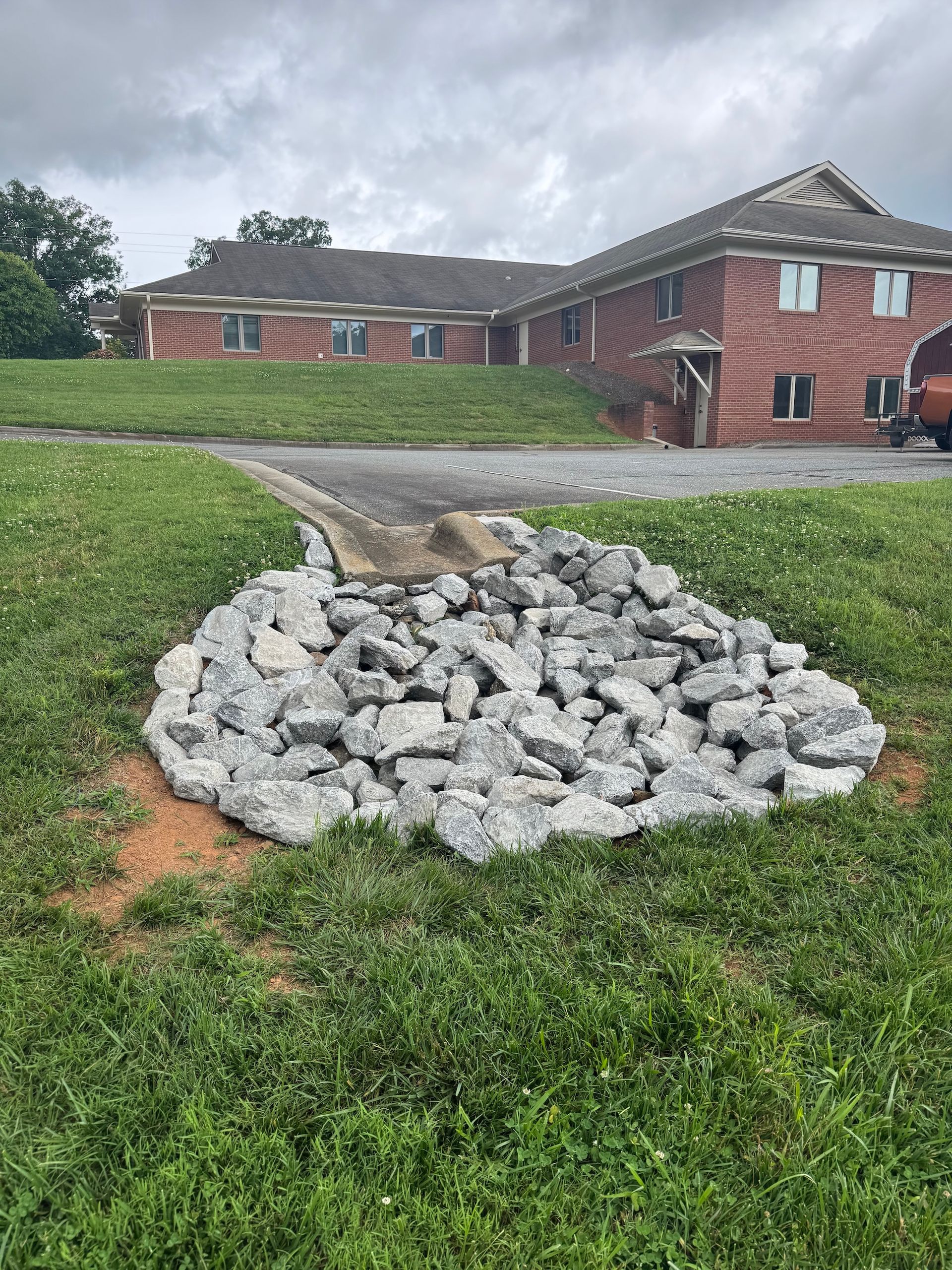 A concrete drainage pipe outlet leading into a bed of grey riprap stones on a grassy lawn in front of a brick building.