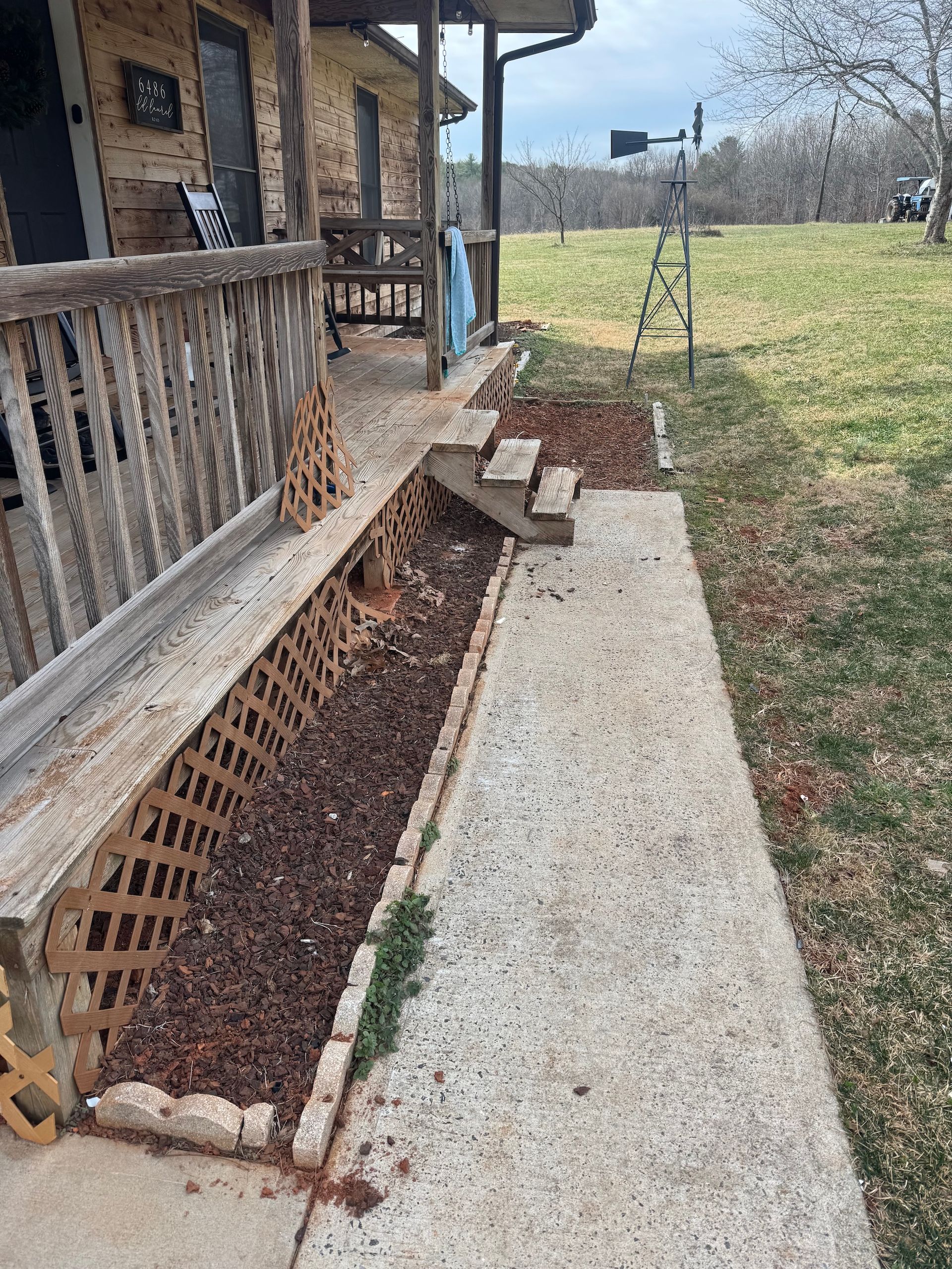 A wooden porch with lattice skirting next to a concrete walkway, leading to a grassy field with a metal windmill.