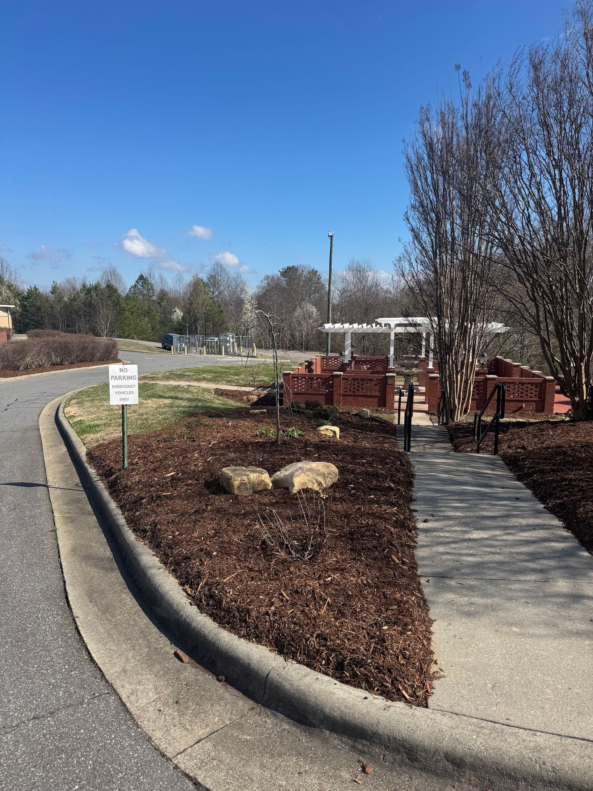 A concrete path curves past a garden with mulch, stones, and a white trellis structure under a clear blue sky.