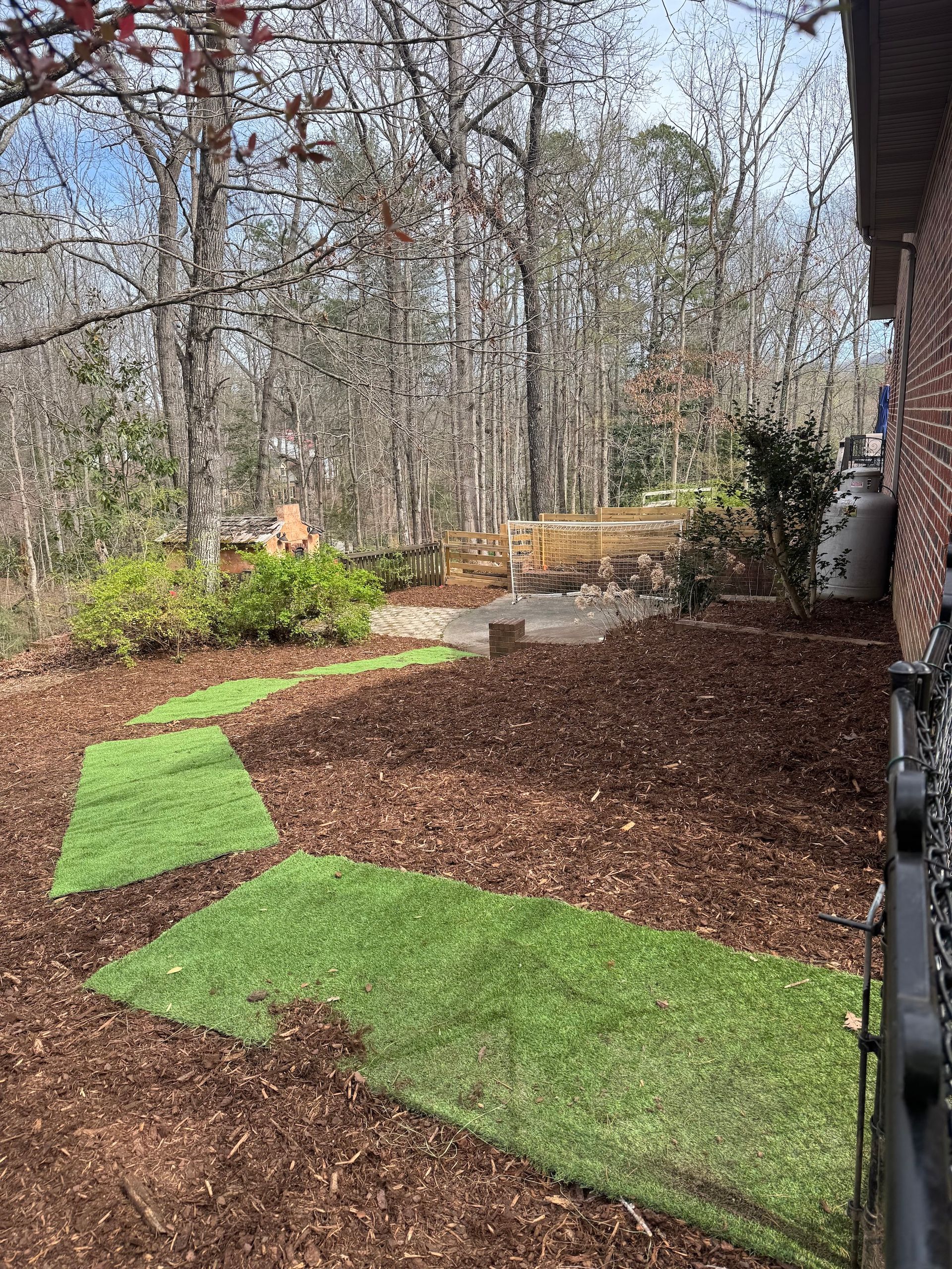 A backyard path made of rectangular artificial grass segments set into a bed of brown wood mulch near a wooded area.