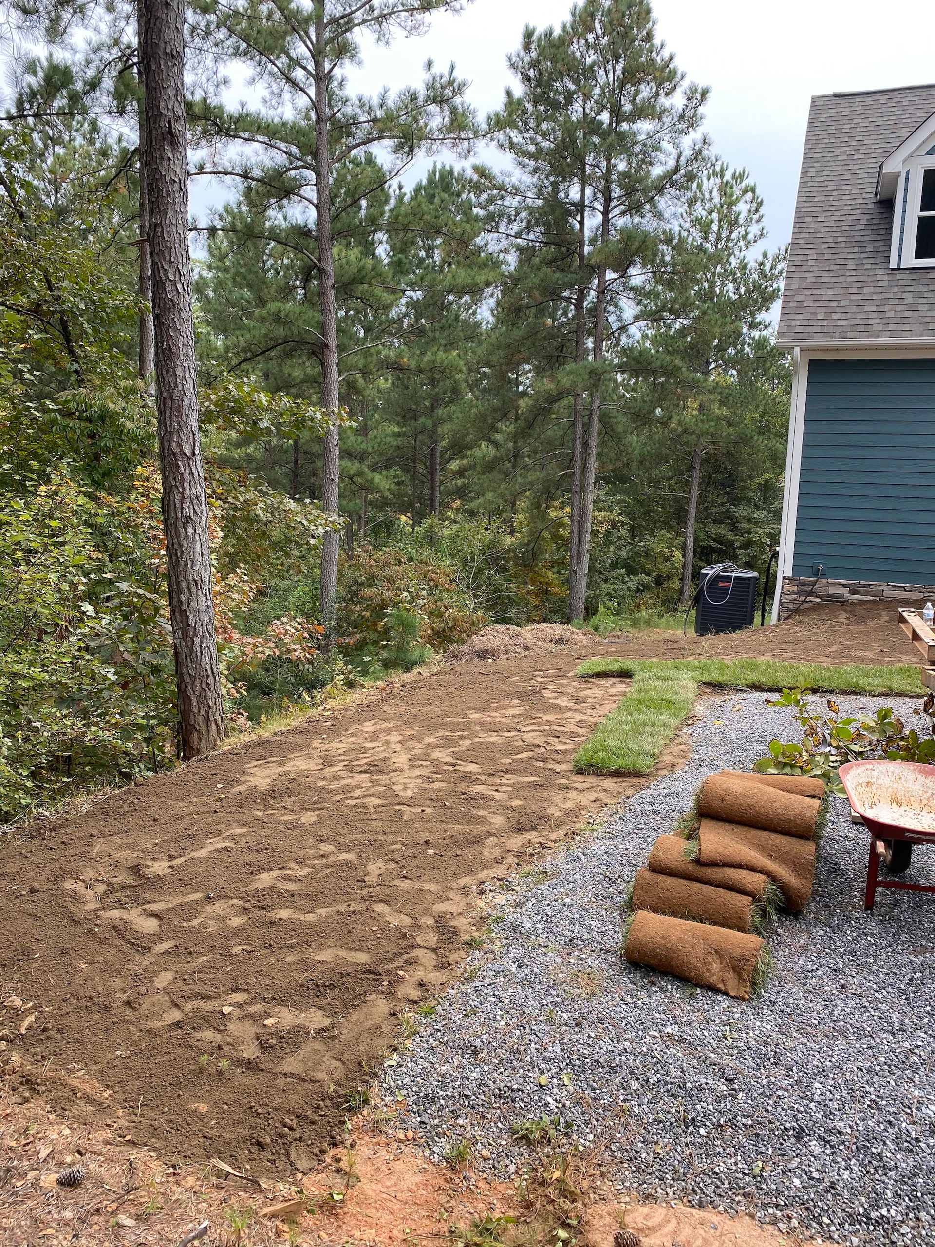 A yard project with rolls of sod waiting to be laid next to a gravel patio and a blue-sided house near a forest.