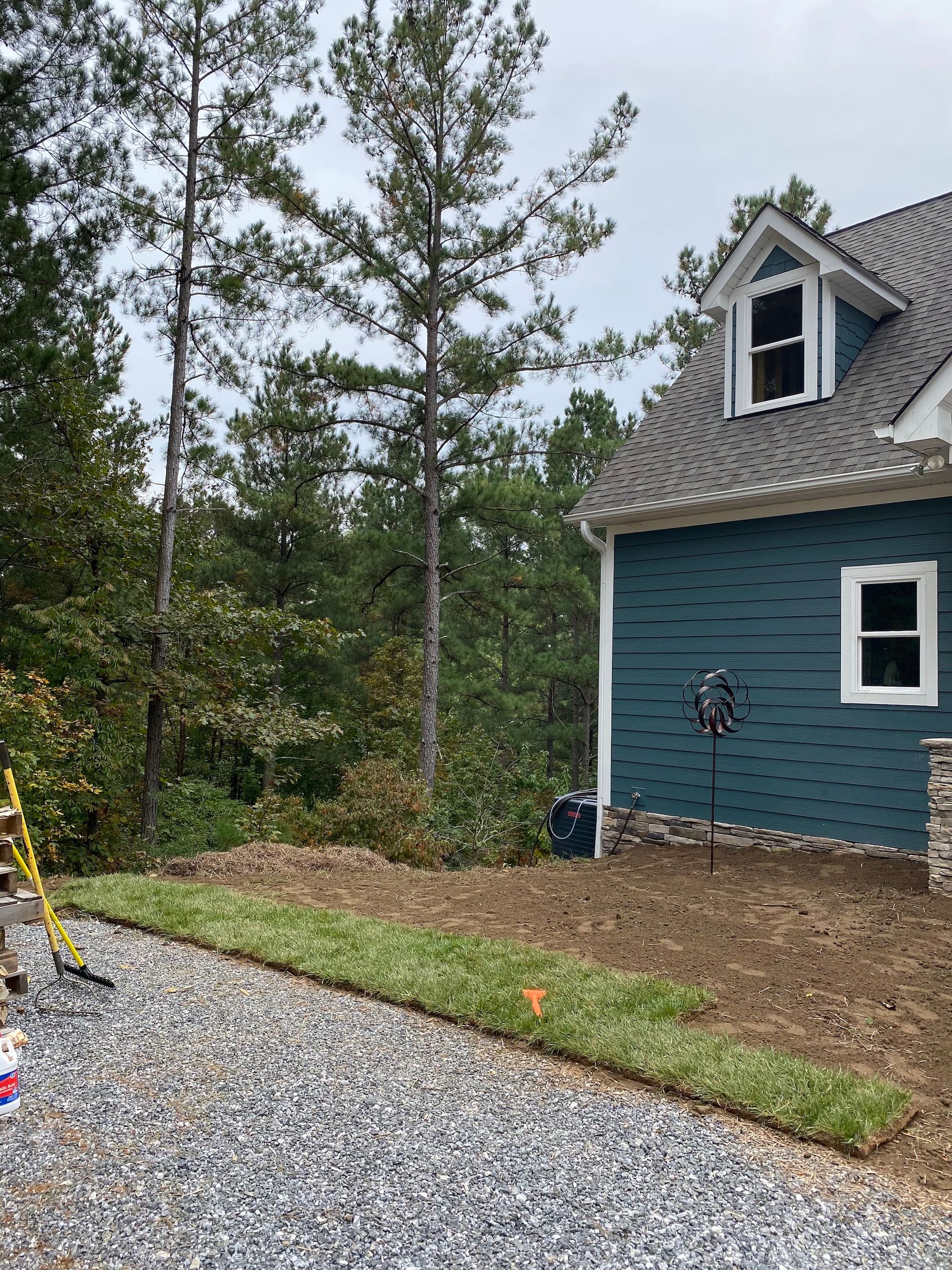 A newly laid strip of green sod runs along a gravel driveway next to a blue house siding with a dormer window.