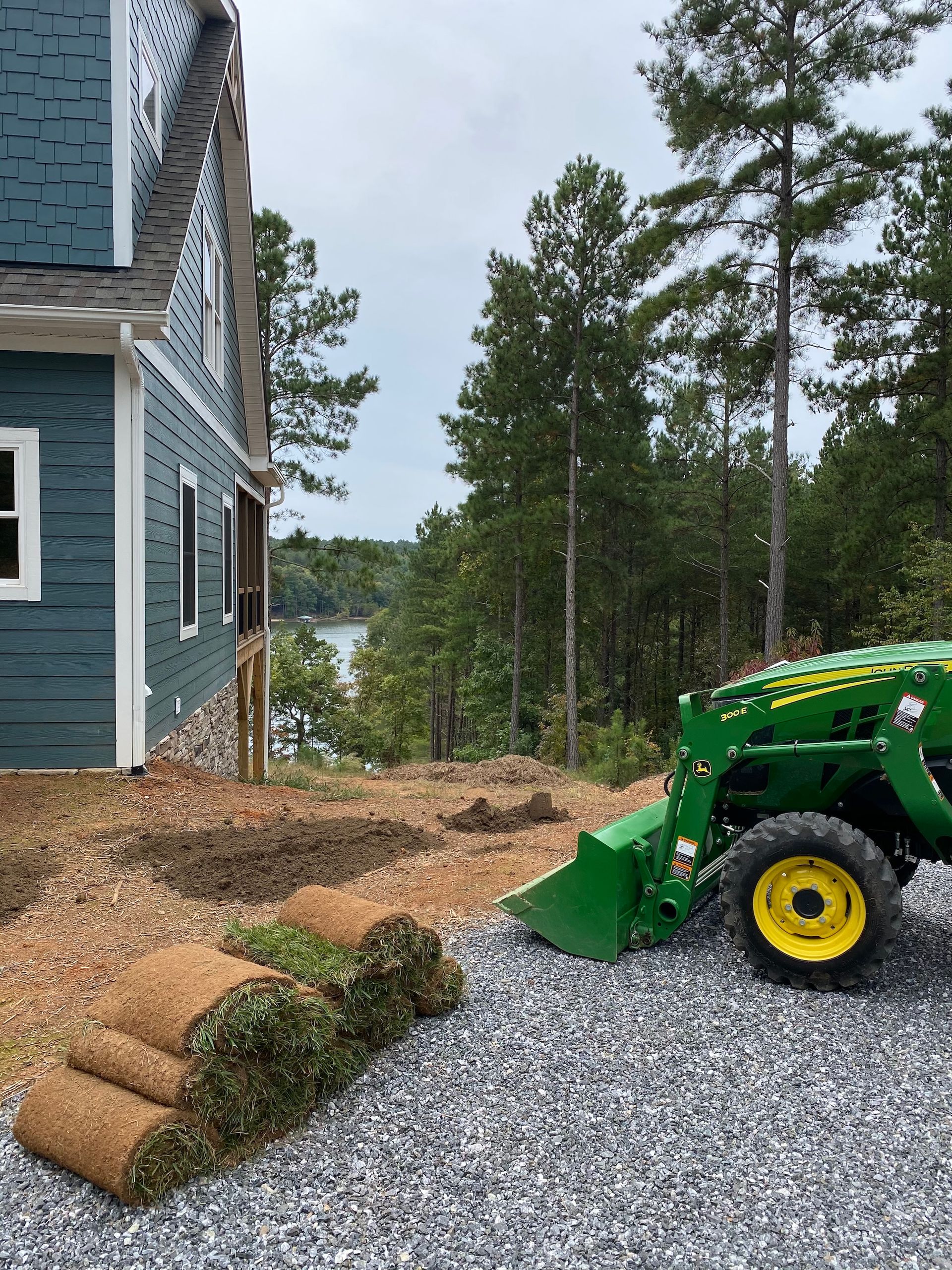A green John Deere tractor sits on a gravel driveway beside a blue house, with stacked rolls of sod and a forest beyond.
