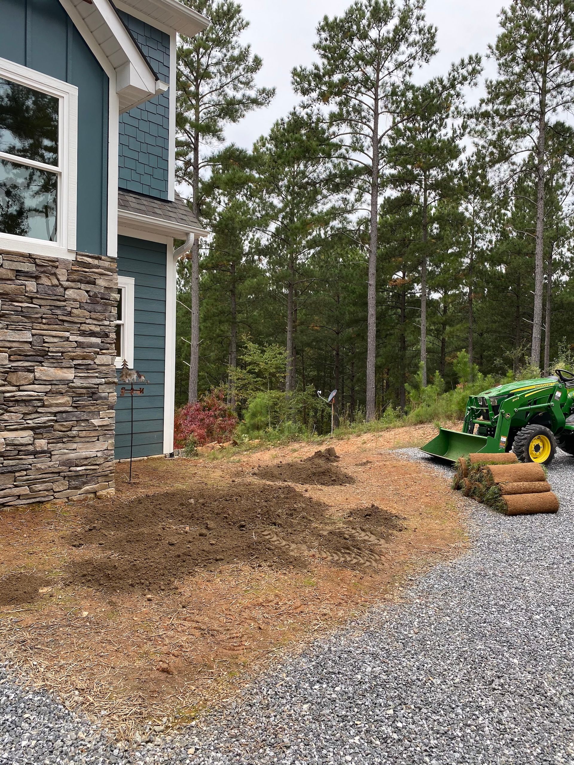 A green lawn tractor sits on a gravel driveway beside a house with blue siding and stone accents, with rolls of sod nearby.