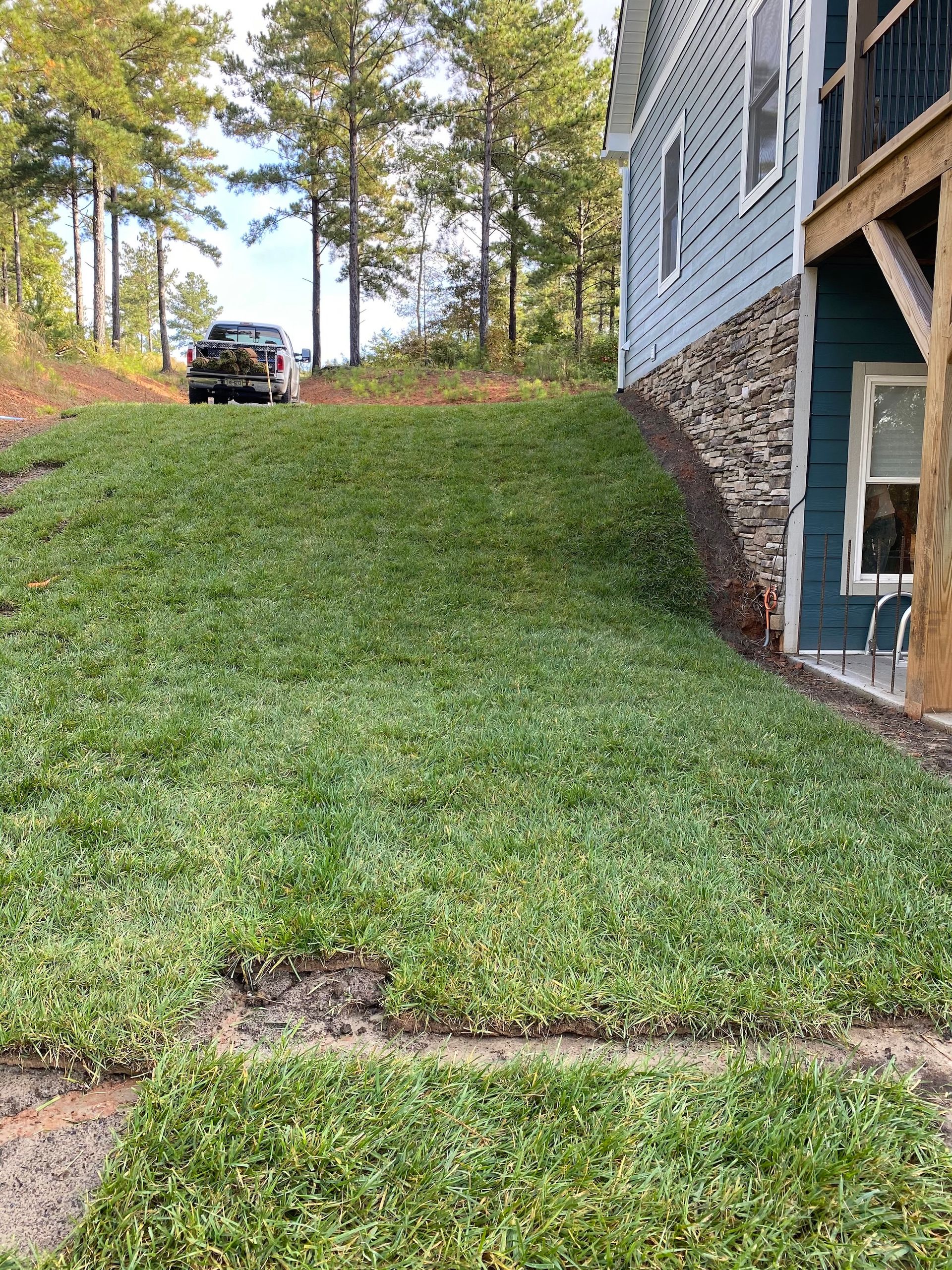 A newly sodded grassy slope runs alongside the stone and blue-sided exterior wall of a house with trees in the background.