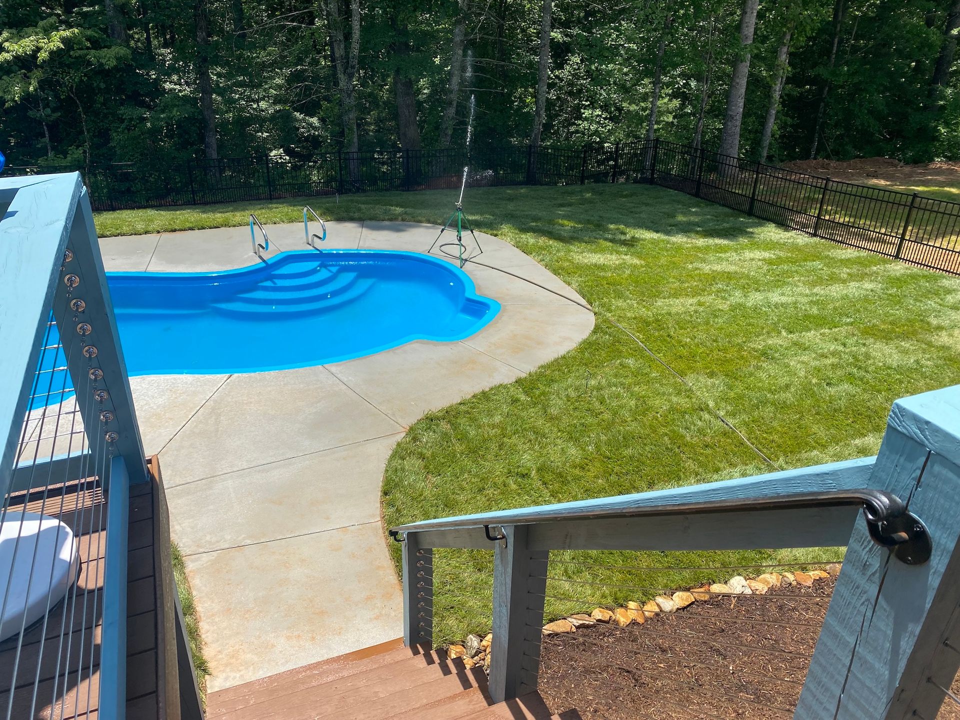 A view from a wooden deck overlooking a bright blue swimming pool, a concrete deck, and a lush green lawn.