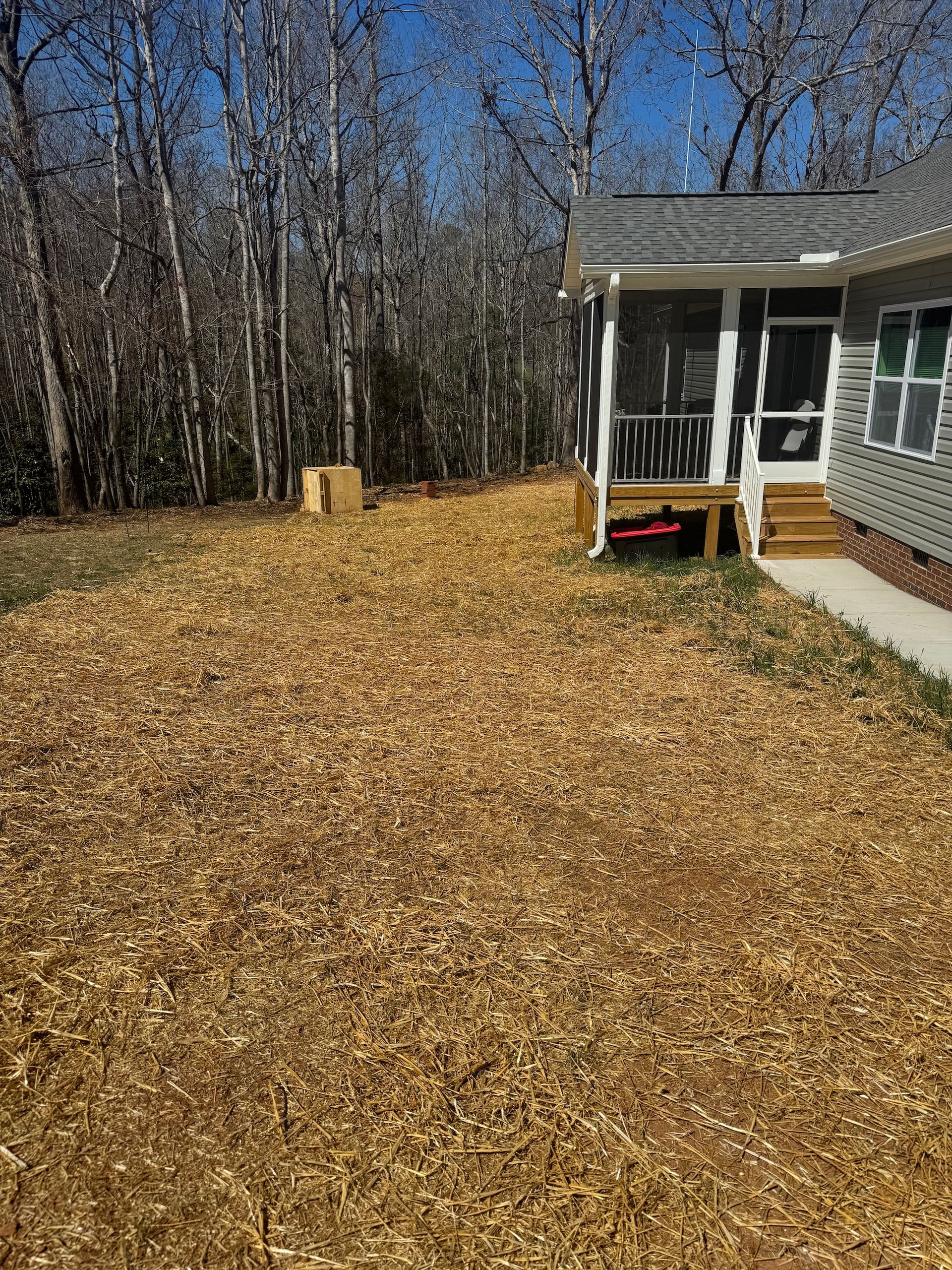 A wooden house with a screened-in porch sits next to a large yard covered in brown wood mulch near a forest.