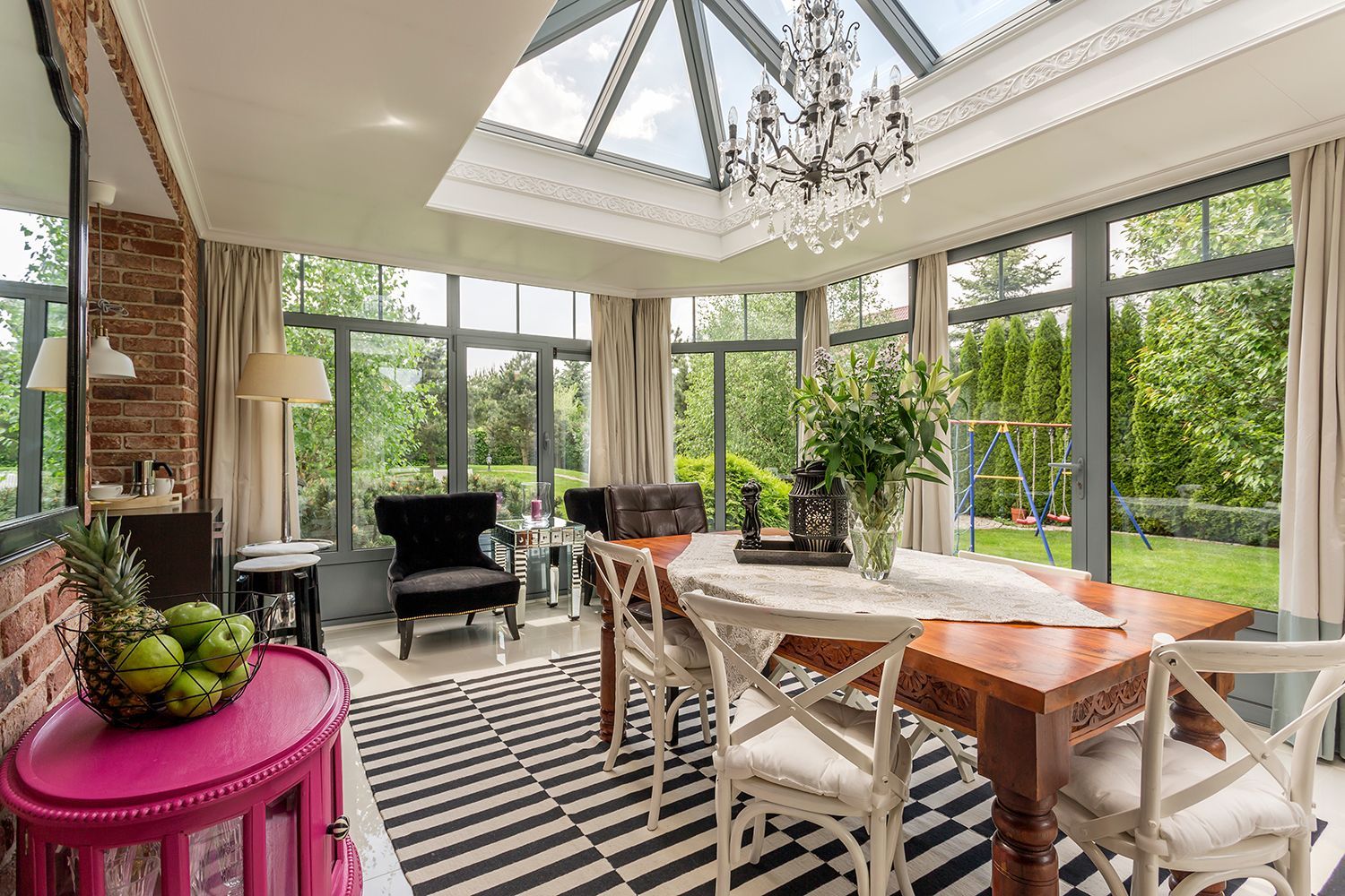 Bright sunroom dining area with large windows, striped rug, wood table, plants, and black chairs Bright sunroom dining area with large windows, striped rug, wood table, plants, and black chairs