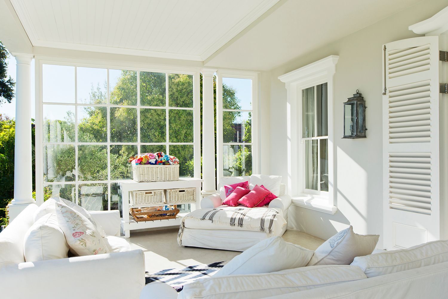 Bright sunroom with white furniture, pink pillows, and large windows overlooking greenery Bright sunroom with white furniture, pink pillows, and large windows overlooking greenery