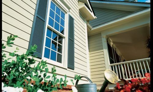 Exterior of a beige house with blue shutters, white balcony railing, and flowers in the foreground