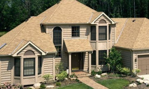 Two-story beige house with tan roof, front porch, and landscaped yard surrounded by trees.