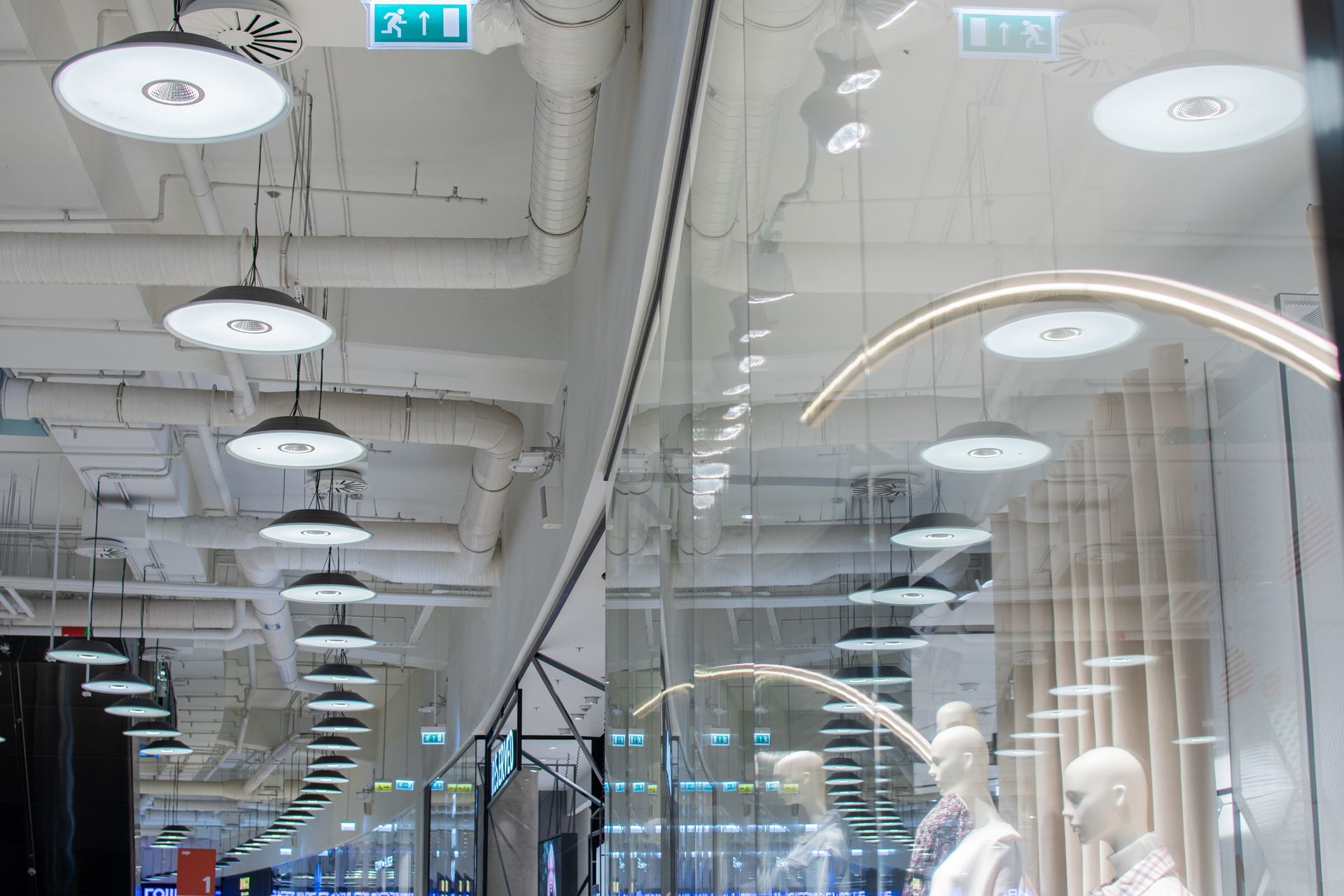 Un plafond de magasin moderne avec des luminaires circulaires blancs suspendus, des conduits de ventilation gris et des panneaux muraux clairs.