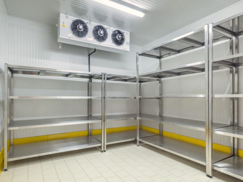 Étagères vides en acier inoxydable dans une chambre froide blanche, plinthes jaunes, ventilateur de plafond.