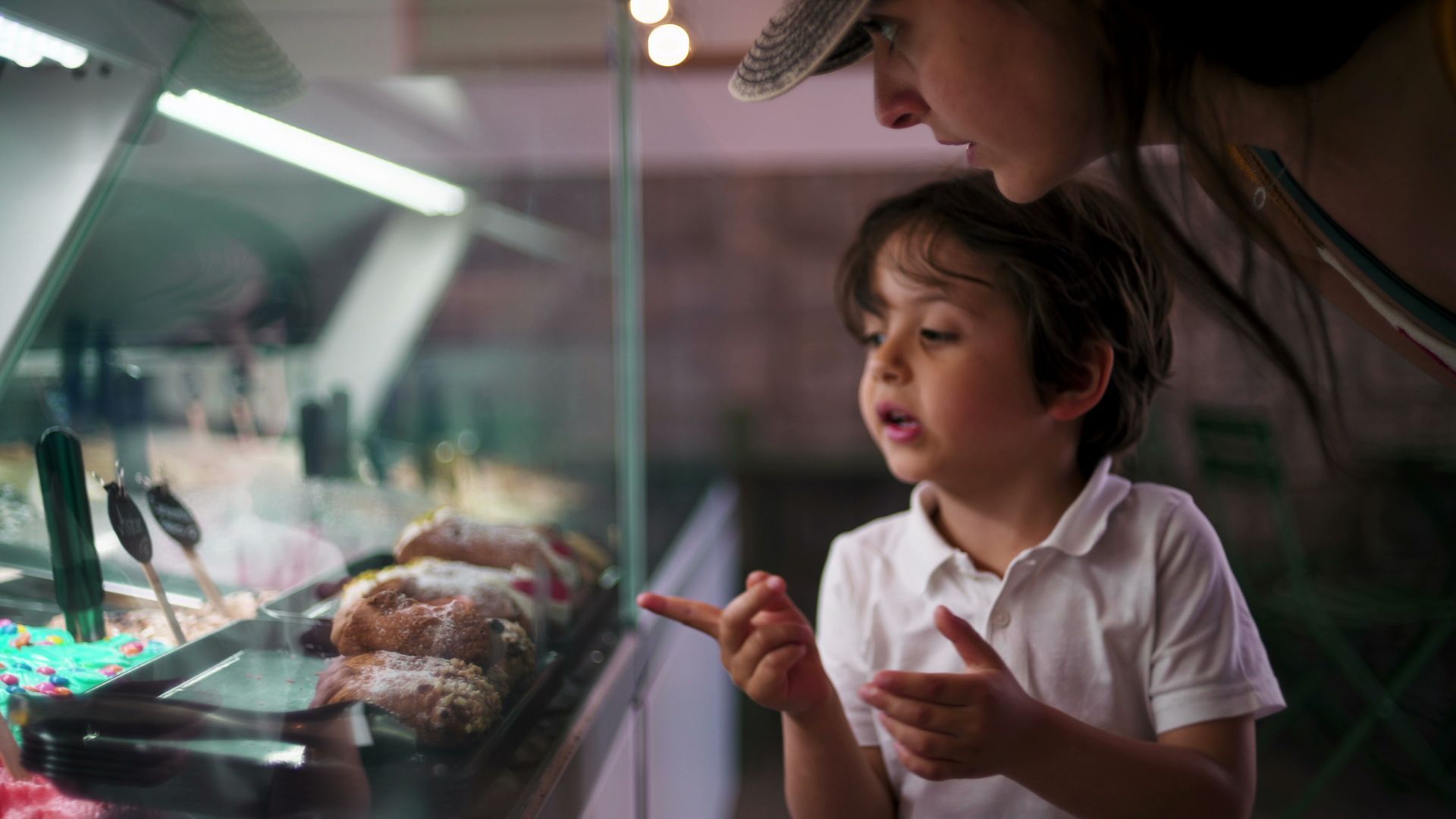 Un garçon désigne du doigt des pâtisseries dans une vitrine, sous le regard d'une personne coiffée d'un chapeau.