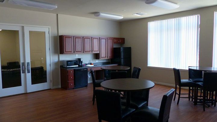 Empty break room with tables, chairs, cabinets, and a kitchenette by a bright window.