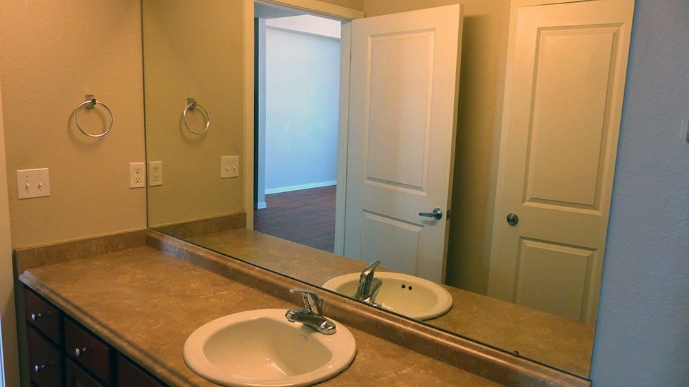 Bathroom vanity with double sinks, mirror, beige walls, and open door to adjacent room