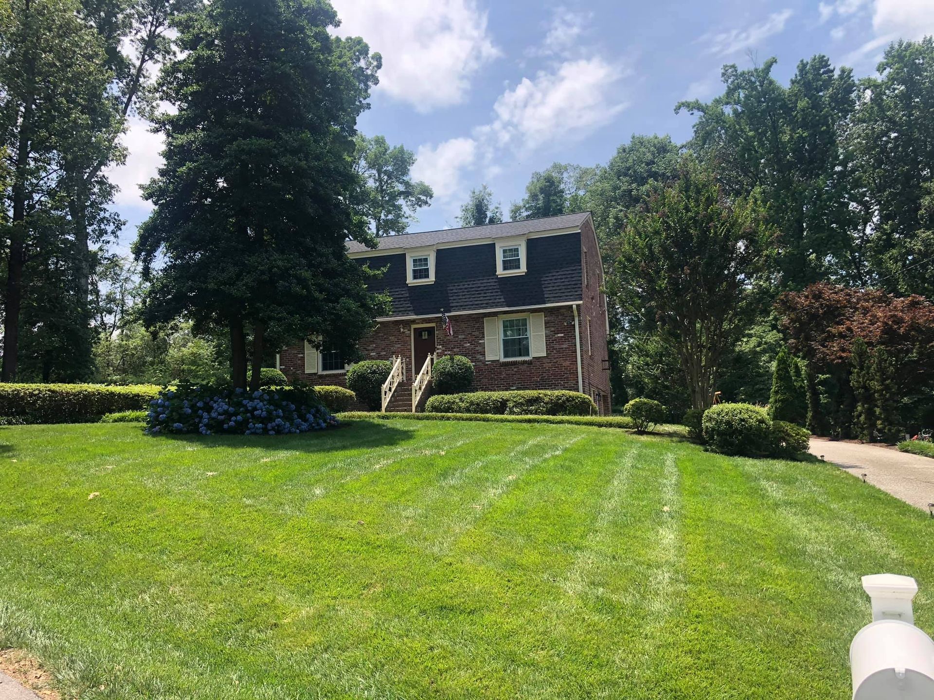 A brick house with a black roof is surrounded by trees and a lush green lawn.