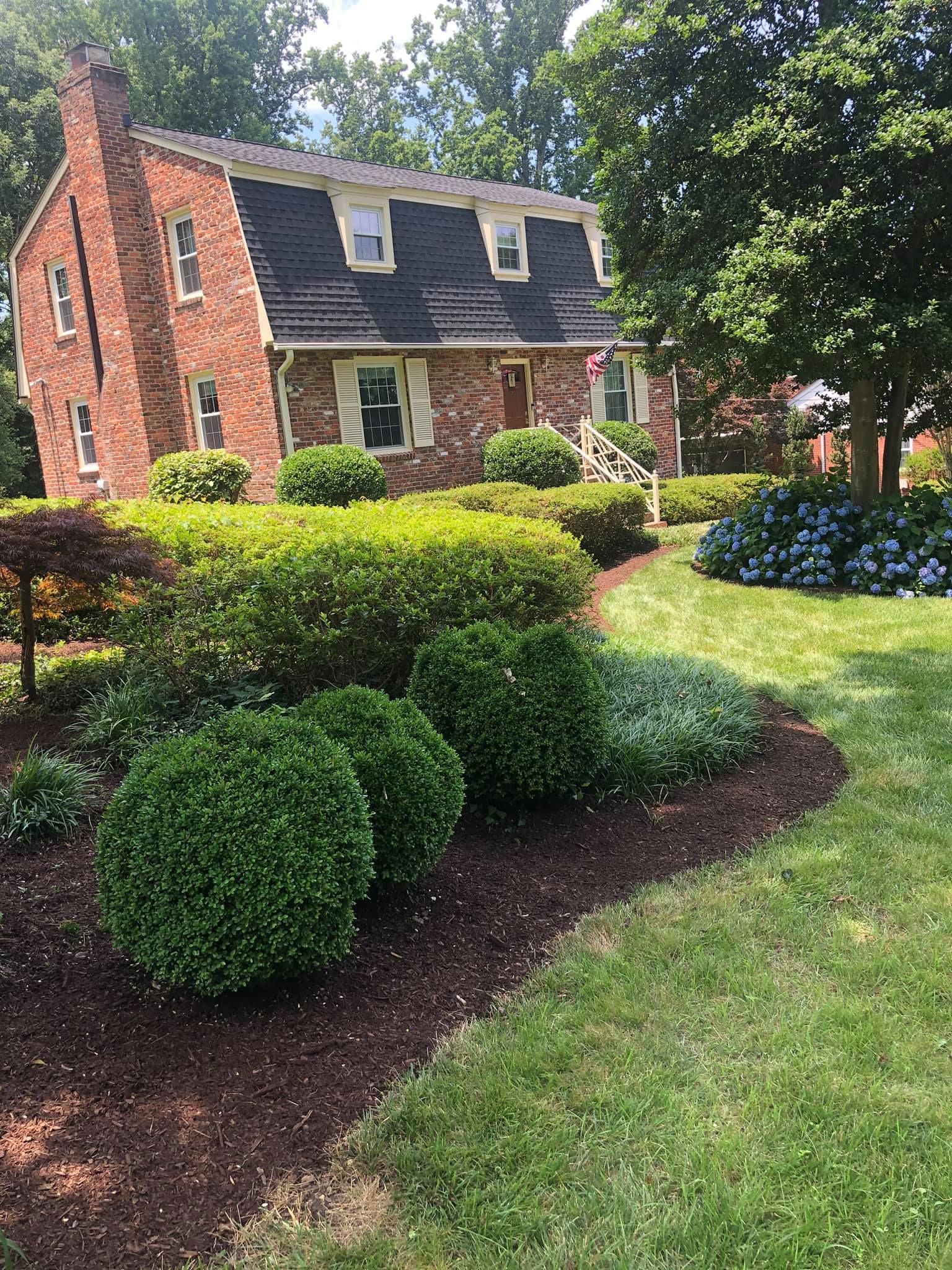 A brick house with a black roof is surrounded by bushes and trees.