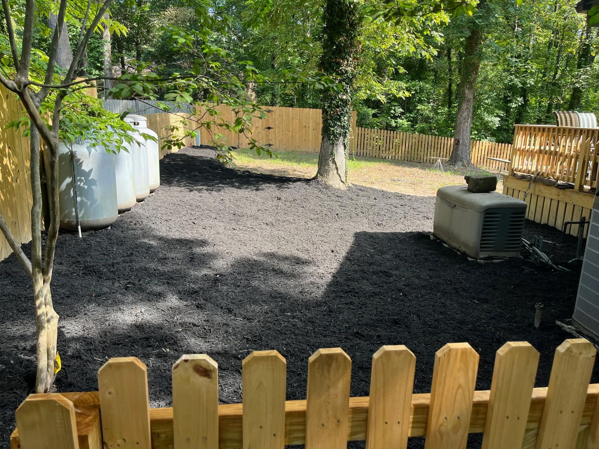A wooden picket fence surrounds a backyard with a propane tank.