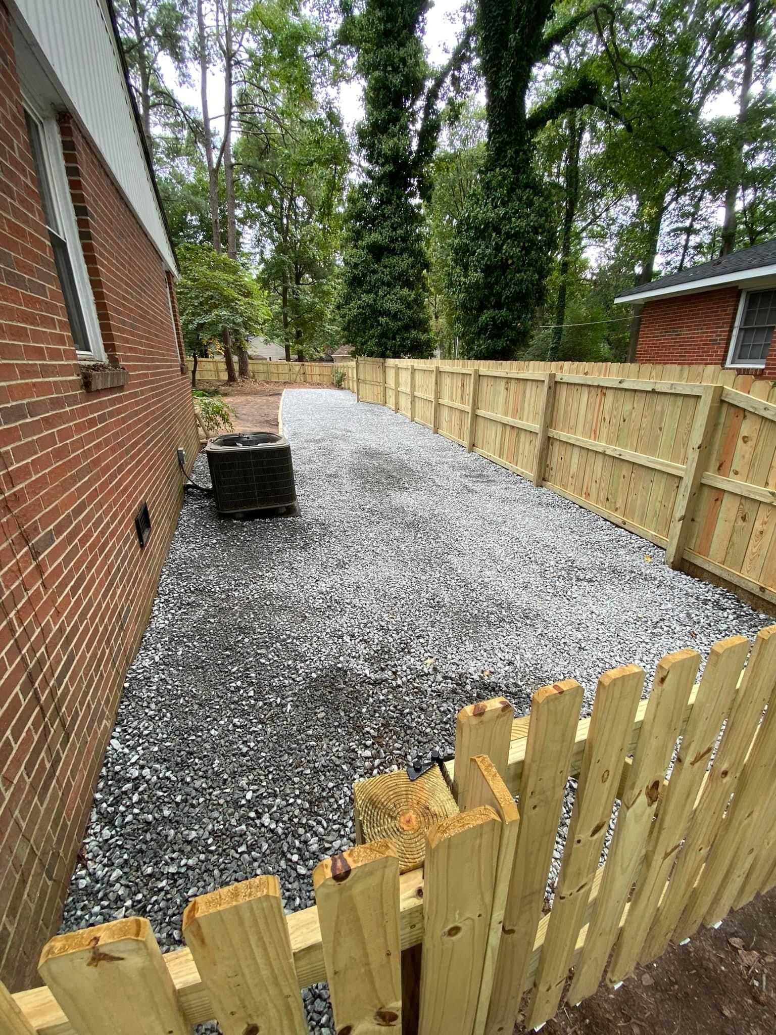 A wooden fence surrounds a gravel driveway in front of a brick house.