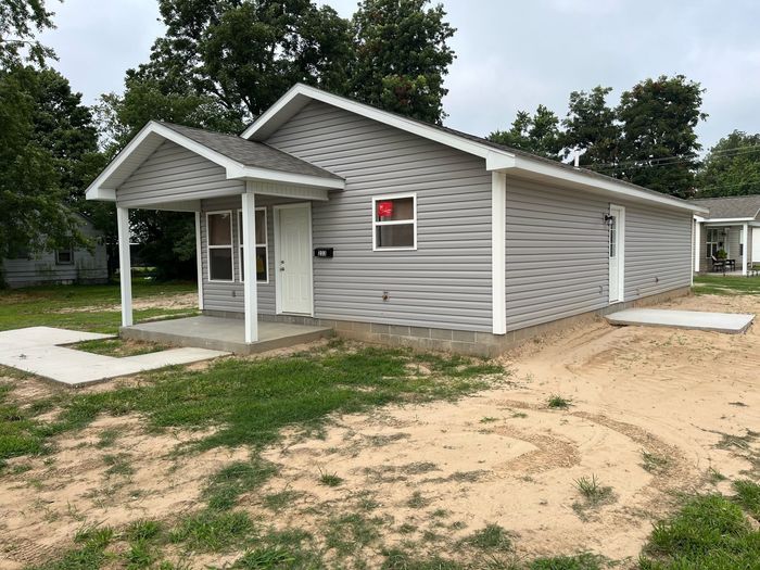 Single-story gray house with a small covered porch and red sign, surrounded by dirt and sparse grass.