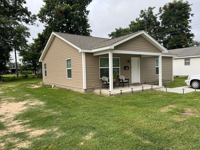 Small beige house with a covered front porch and white trim, surrounded by a grassy yard
