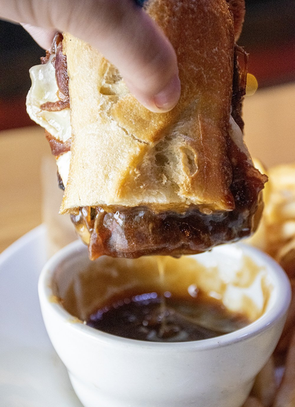 Hand dipping a sandwich into a bowl of dark dipping sauce. The sandwich is on a crusty roll, filled with meat and cheese.